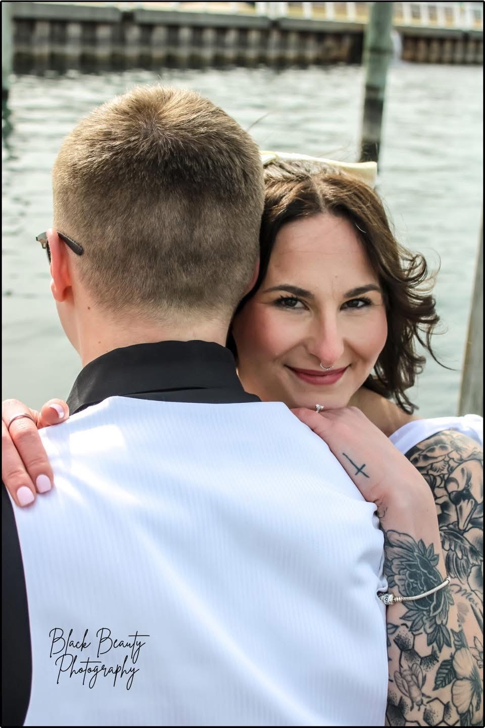 Woman with dark hair and tattoos smiling, hugging a person with short hair and glasses near water.