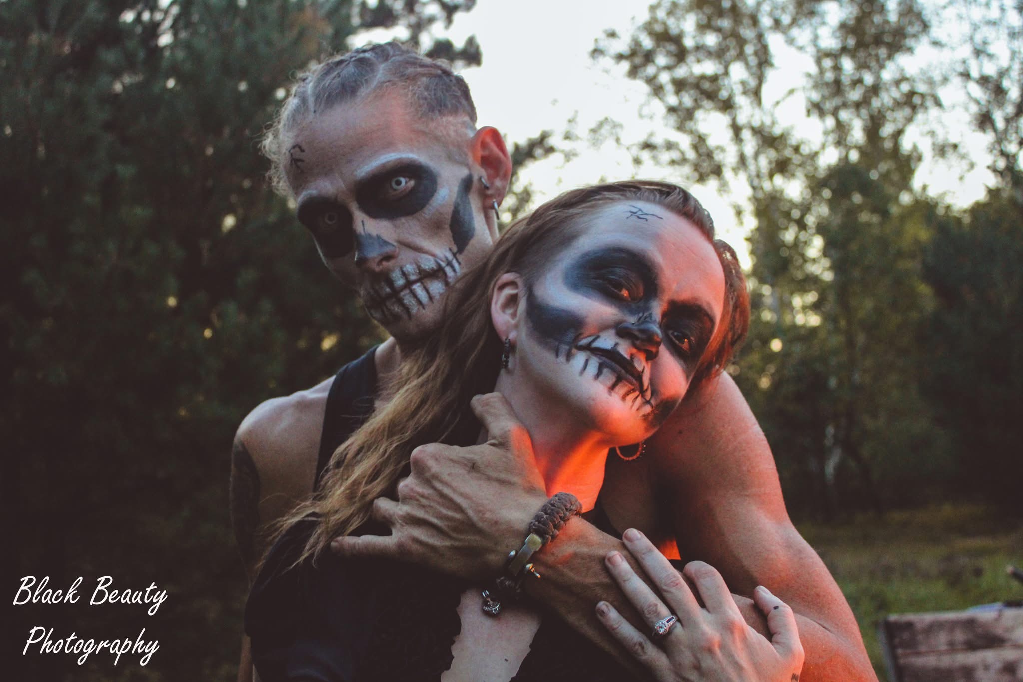 Two people with skull face paint and dark makeup pose outdoors, one holding the other's neck, trees in background.