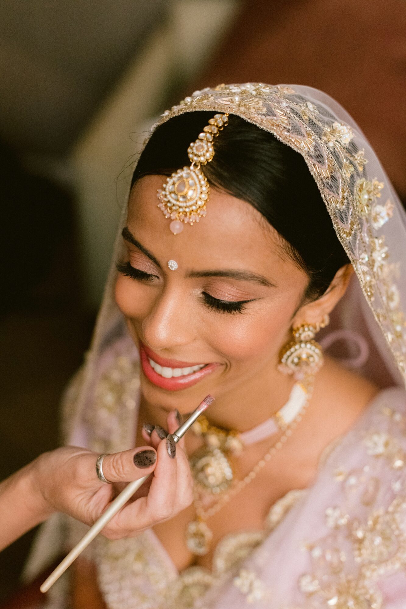 Close-up of a woman with traditional jewelry and a veil, smiling as makeup is applied.