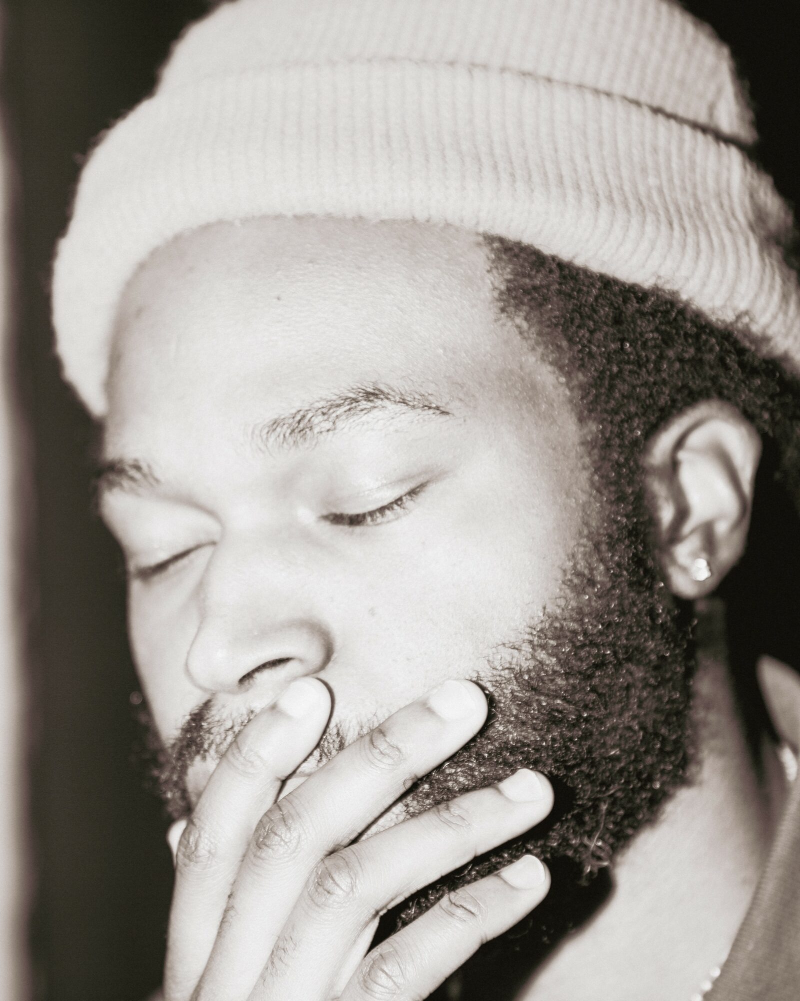 Person with eyes closed, wearing a beanie, touching face with fingers, beard visible, black and white photo.