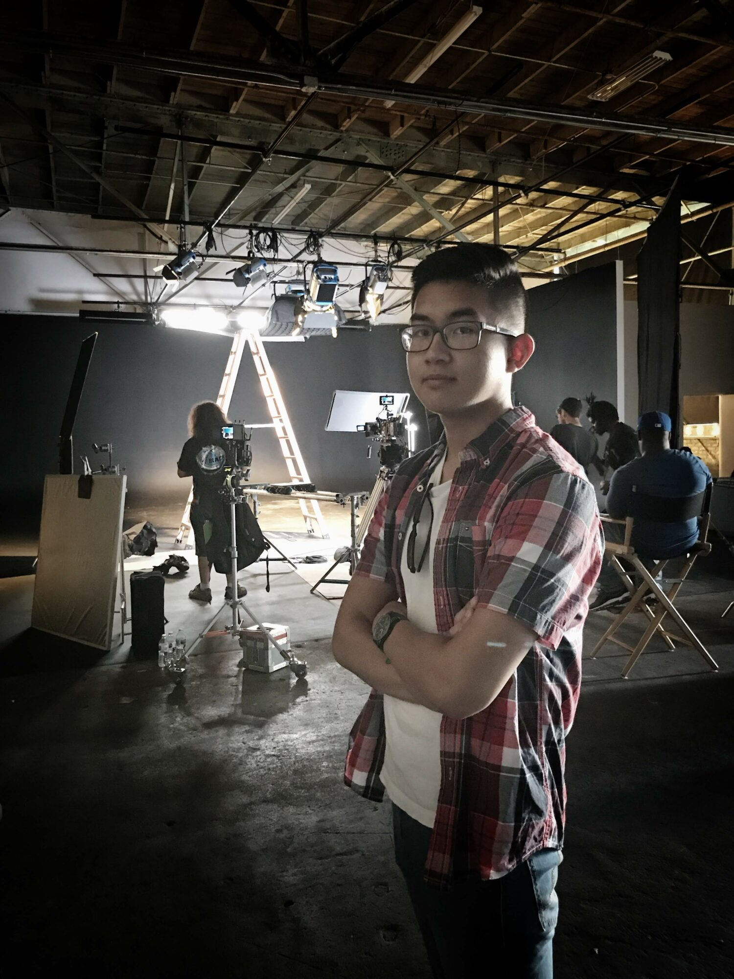 Young man with glasses and plaid shirt standing with arms crossed in a studio or warehouse setting, with people and equipment in the background.