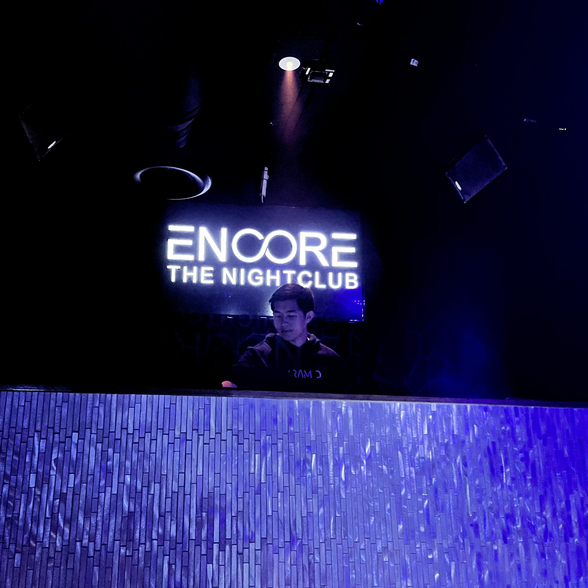 Person standing behind a DJ booth at a nightclub with a sign reading 'ENCORE THE NIGHTCLUB' in the background.