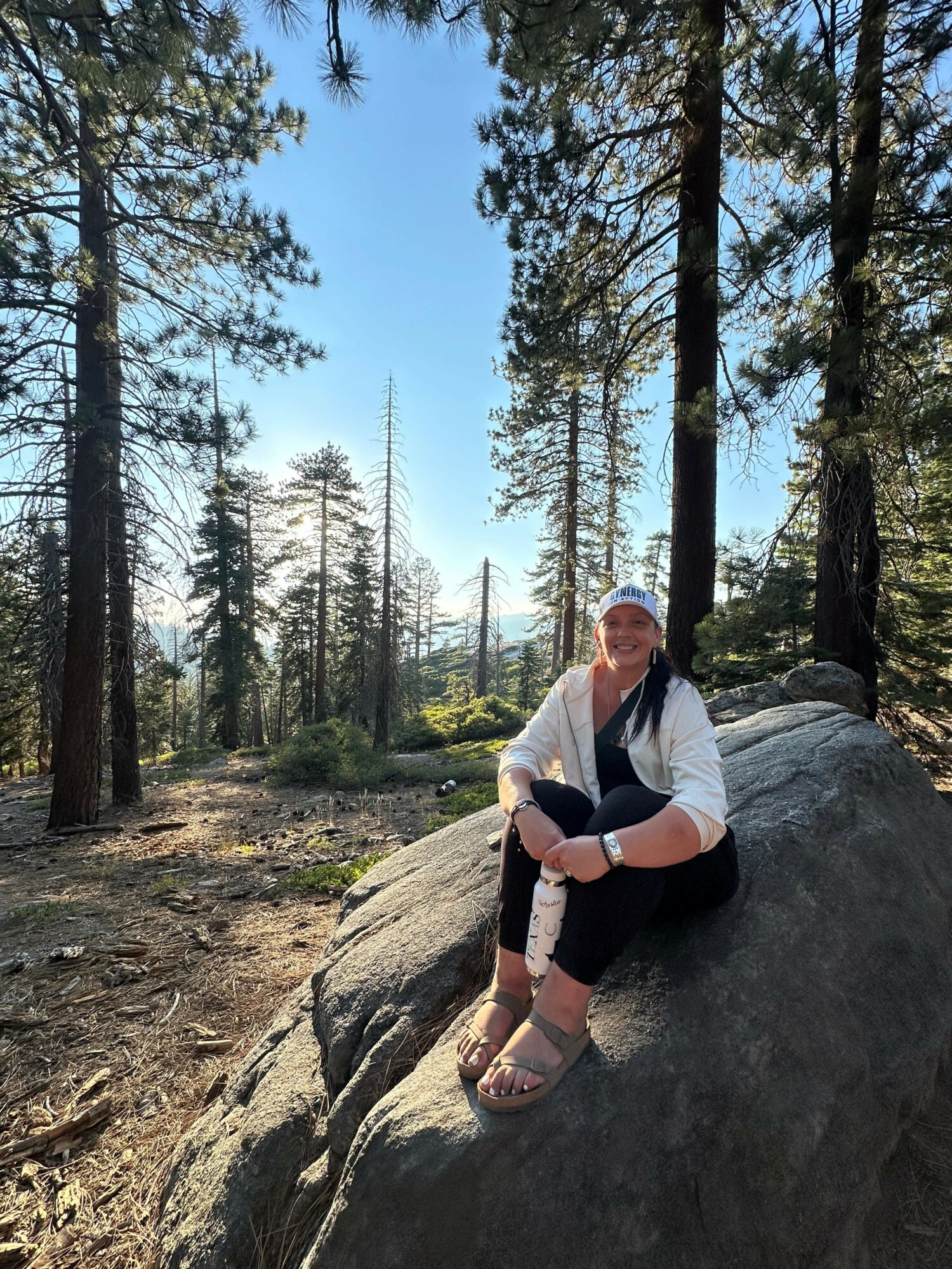 Person sitting on a large rock in a forest with tall trees and blue sky, smiling, wearing a white shirt, black pants, and sandals.