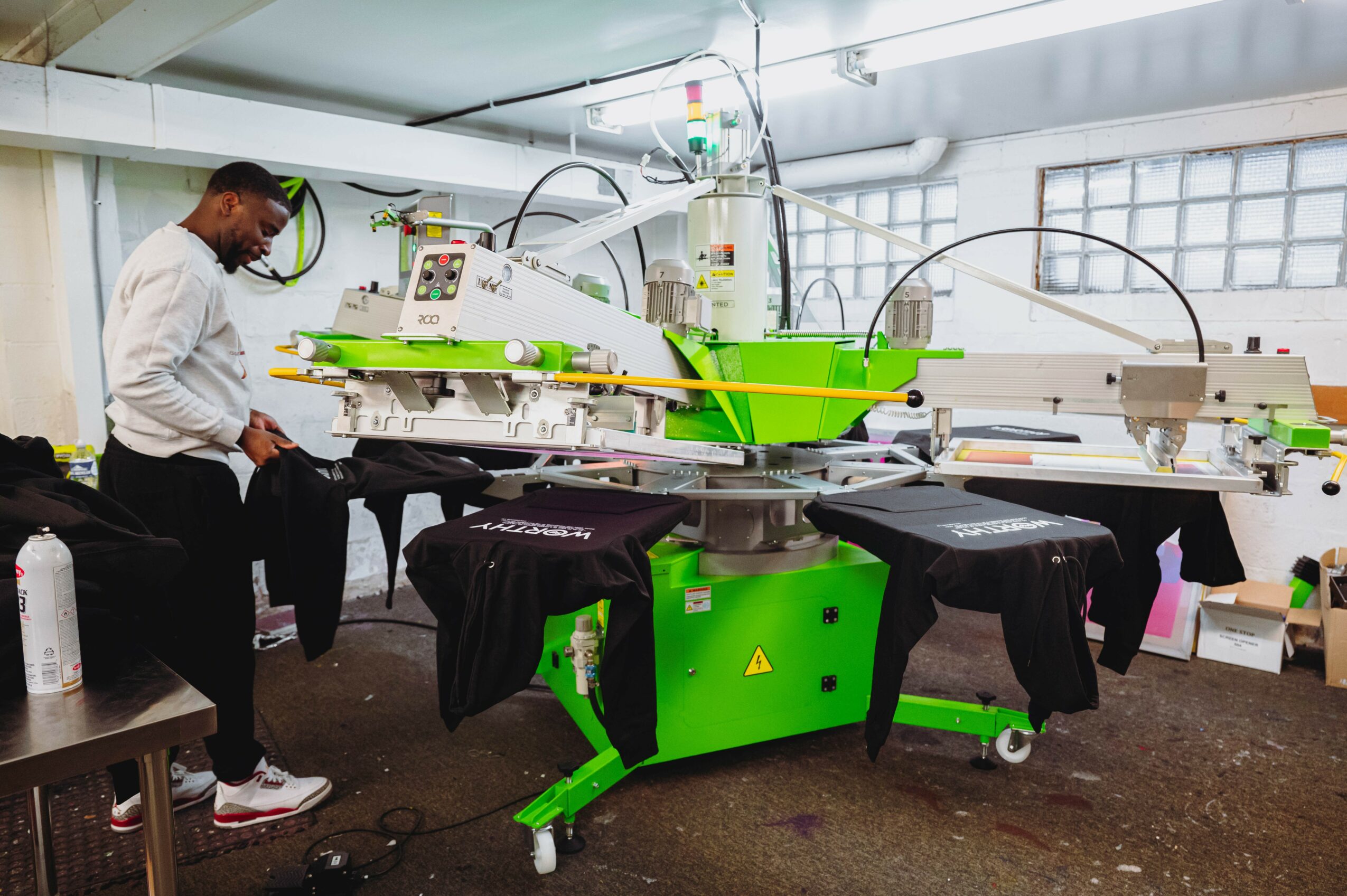Person working with a large, green and white industrial machine in a room with windows, black fabric on the machine, and a table nearby.