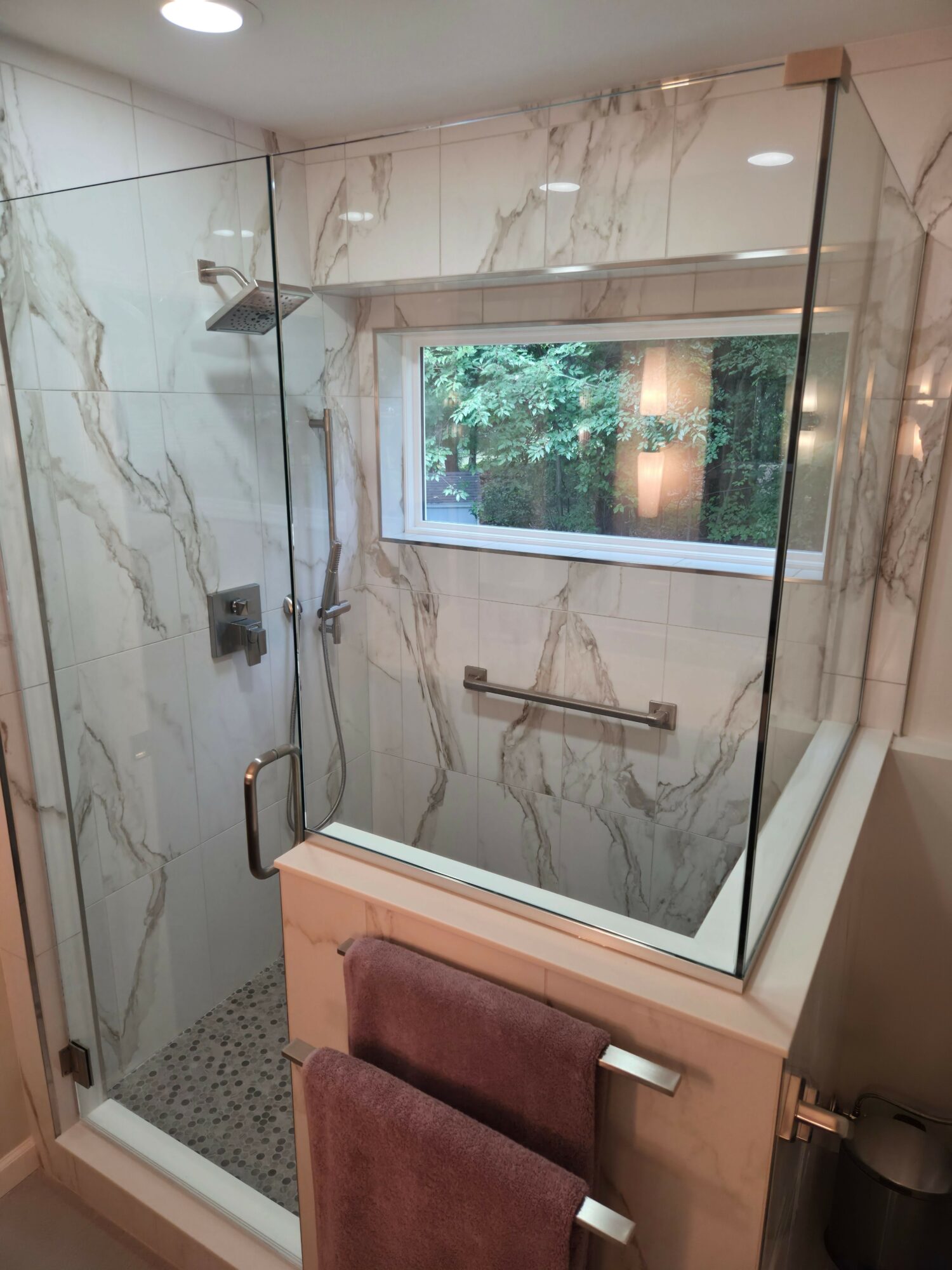Bathroom shower with glass enclosure, marble walls, and a window showing greenery outside.