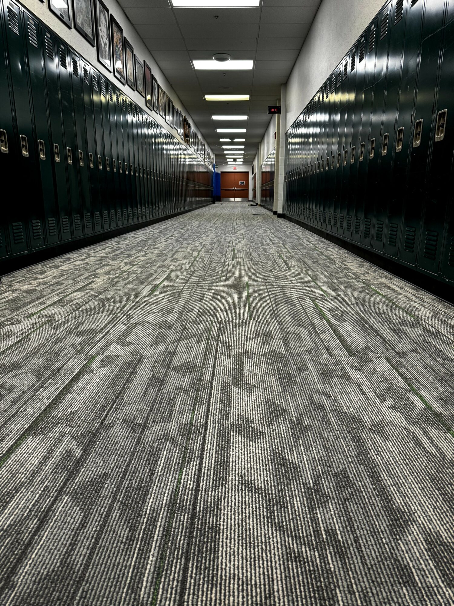 School hallway with green lockers on both sides and a carpeted floor, leading to a distant door.