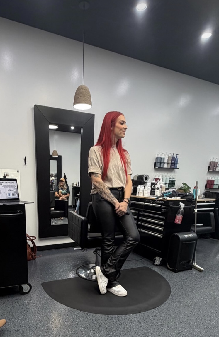 Woman with red hair standing in a modern salon, surrounded by mirrors, chairs, and hair styling tools.