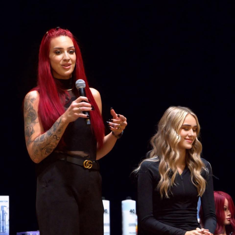 Two women on stage, one with long red hair holding a microphone, the other with long blonde hair smiling.
