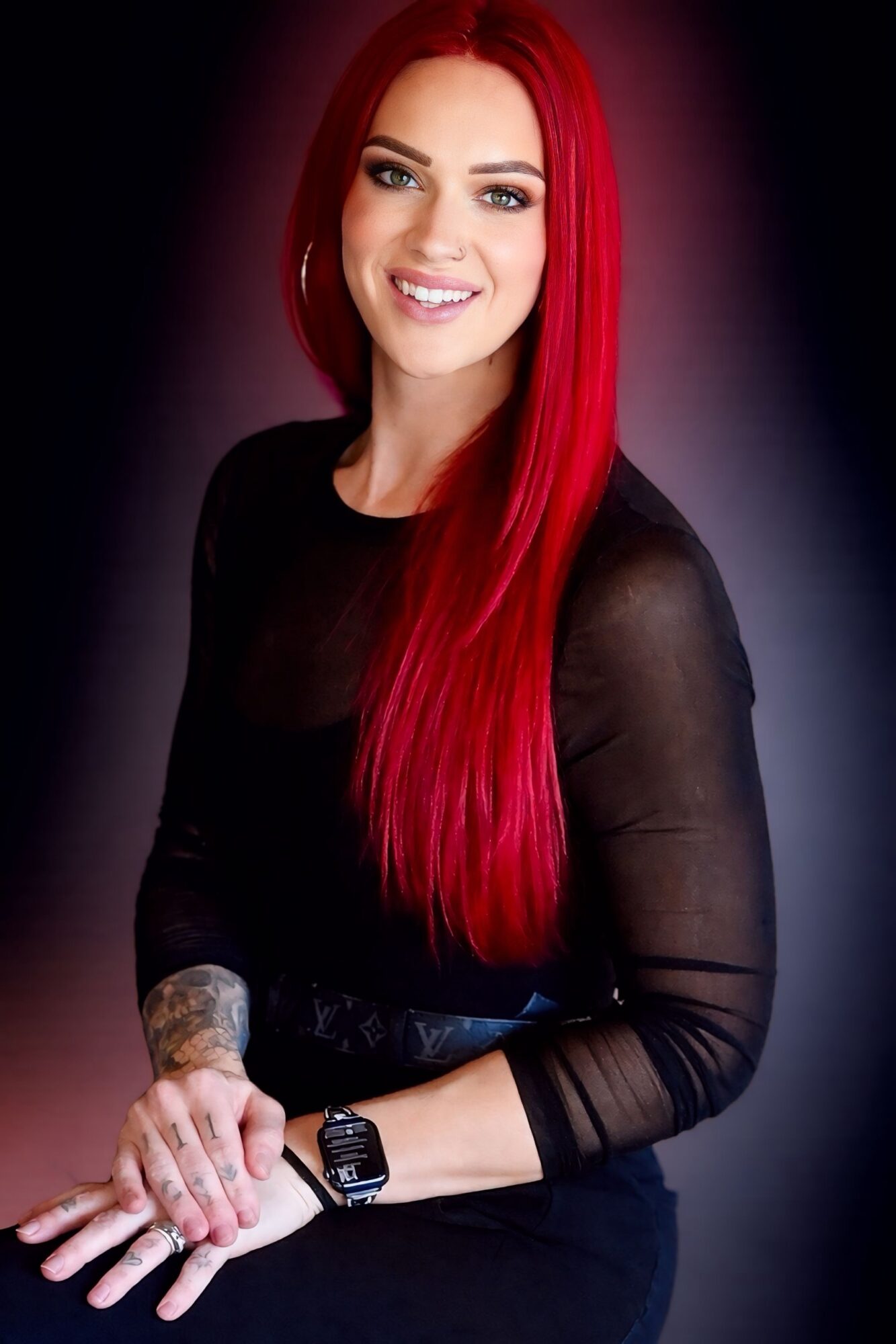 Woman with long red hair smiling, wearing a black sheer top, sitting with hands clasped, dark background