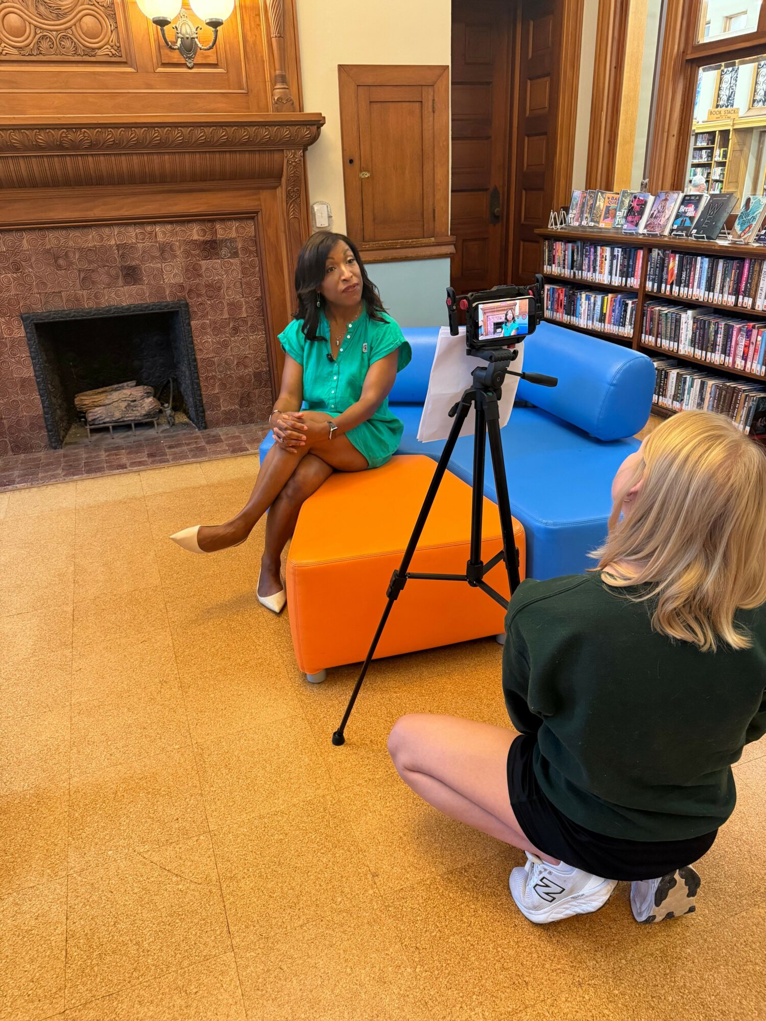 Two children sitting on the floor in a room with a fireplace and bookshelves, one using a camera on a tripod.
