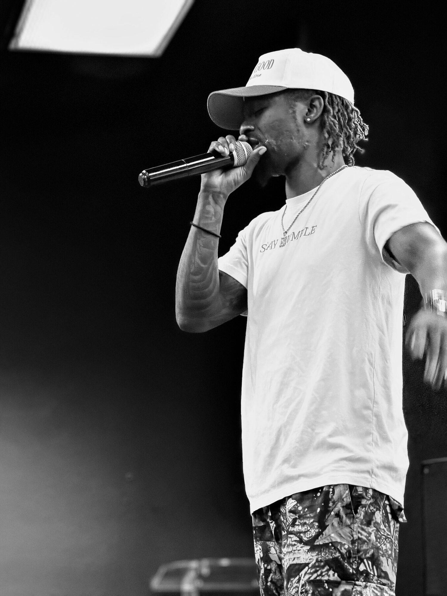 Person performing with a microphone, wearing a cap and casual clothing, on stage.