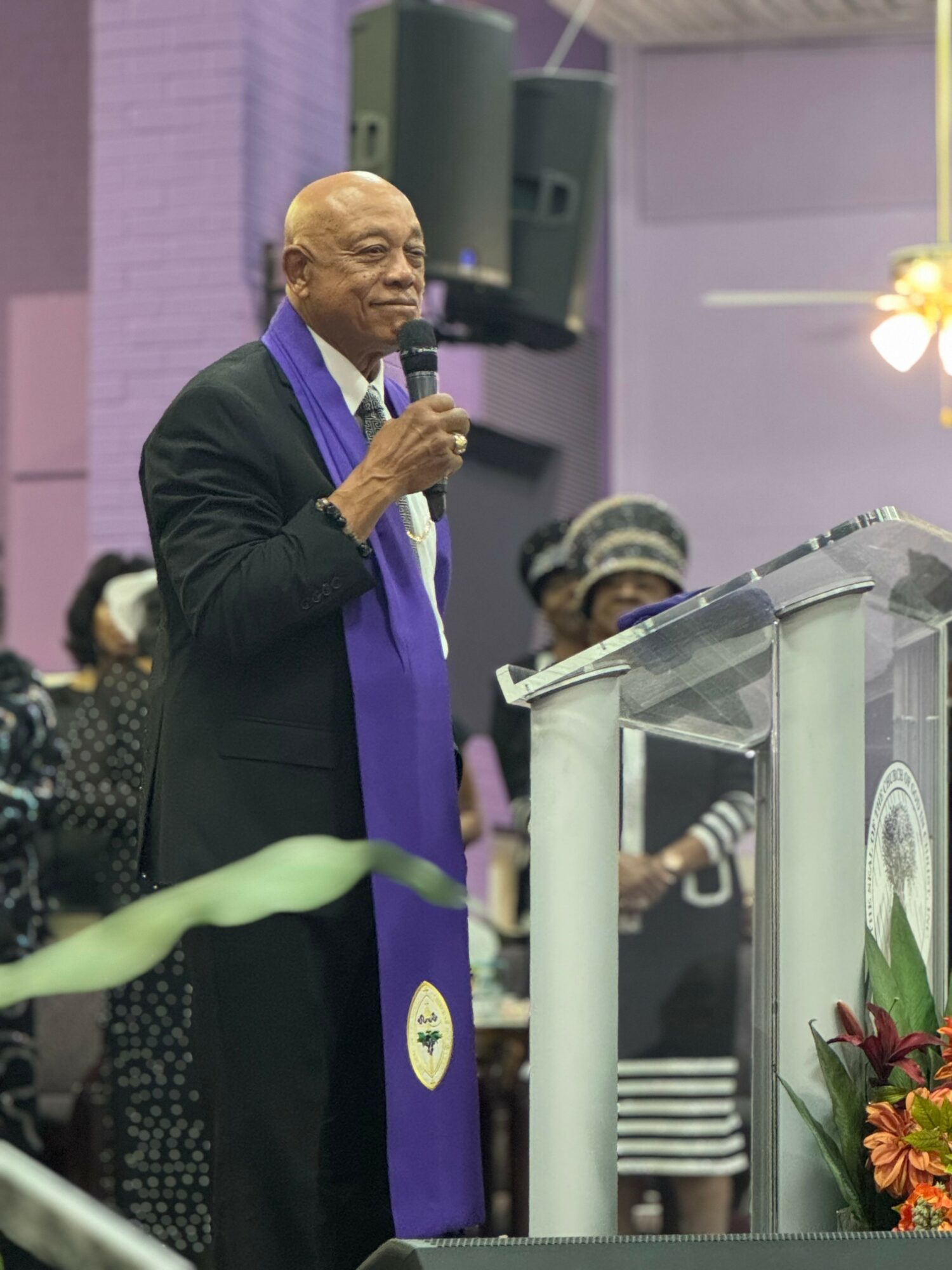 Elderly man speaking at a podium with a microphone, wearing a black suit and purple scarf, in a decorated indoor setting.