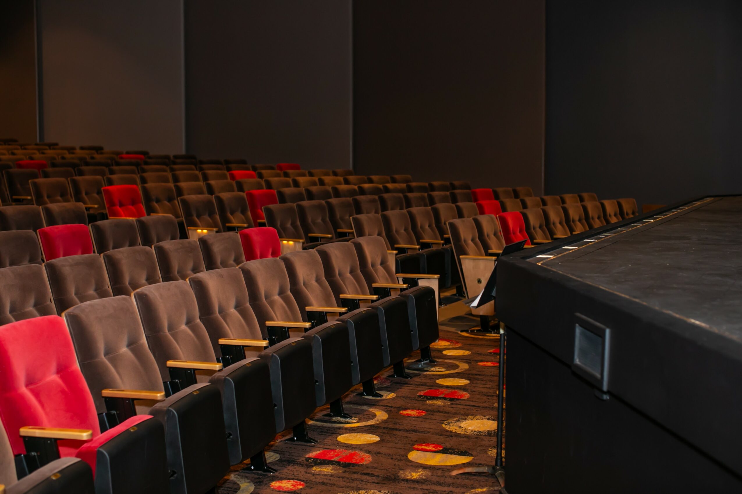 Empty theater with rows of brown and red seats, stage on the right, dark walls, and patterned carpet.