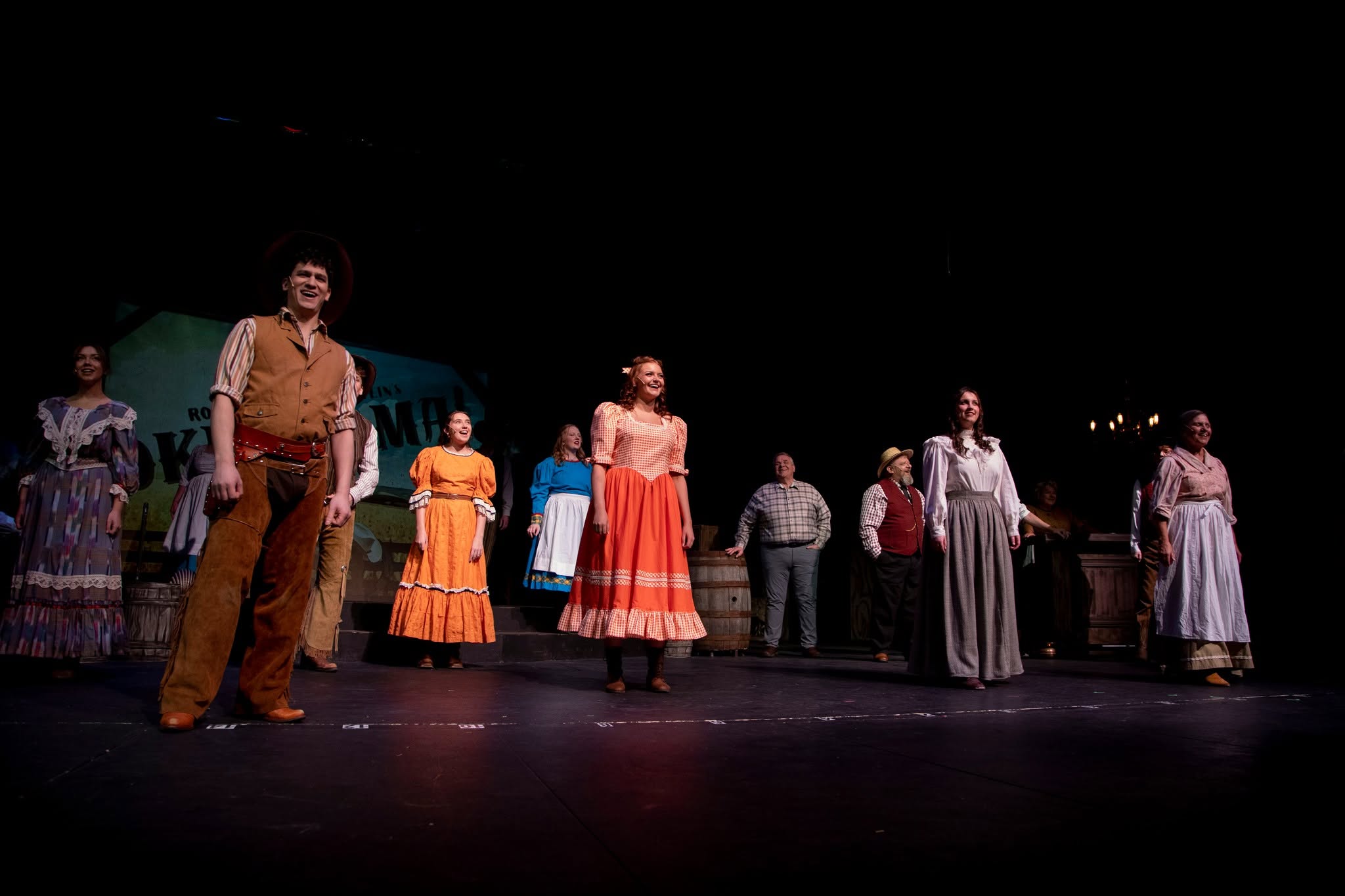 Group of performers on stage, some in colorful costumes, standing in a line, with a dark background.