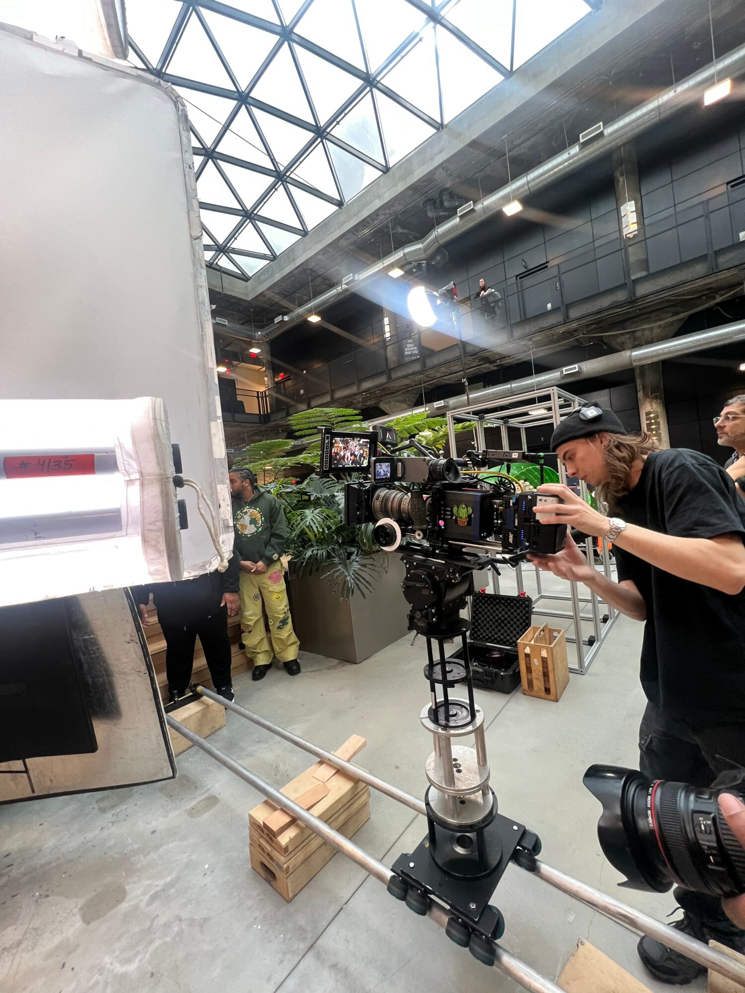 A person operating a camera on a tripod in a modern indoor space with a glass ceiling and people in the background.