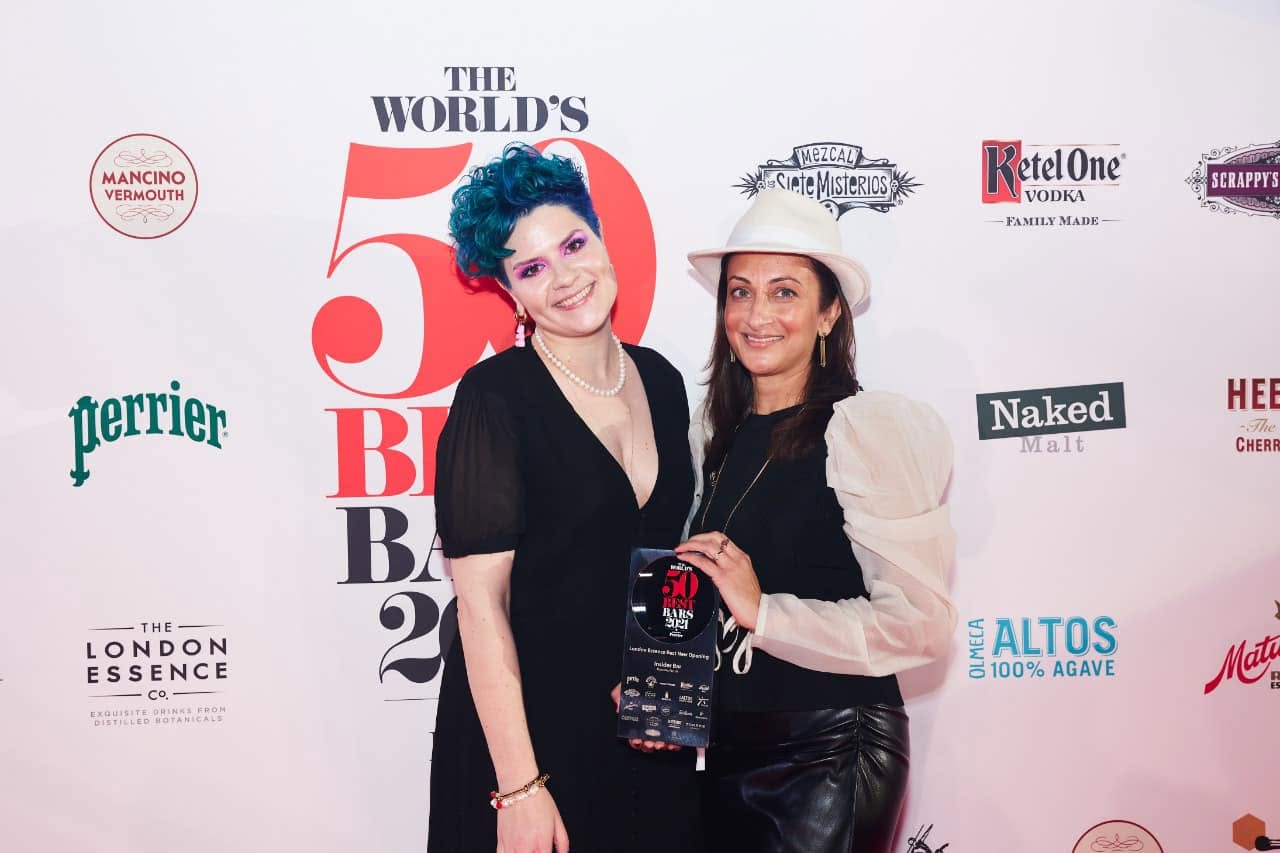 Two women standing in front of a backdrop with logos and text, one with blue hair and the other wearing a hat, smiling.