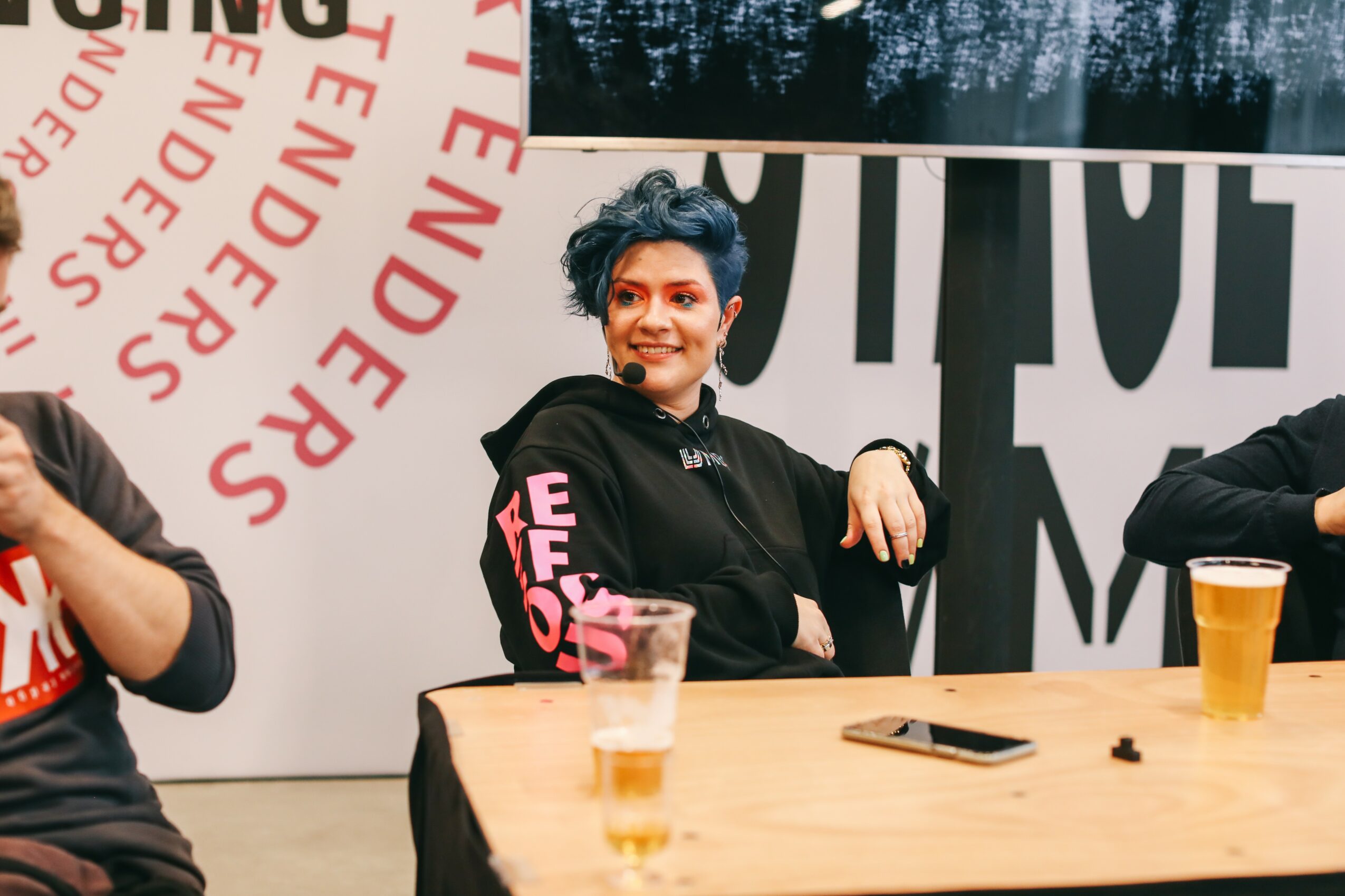 Woman with short dark hair and a black hoodie sitting at a table with drinks and a phone, smiling.