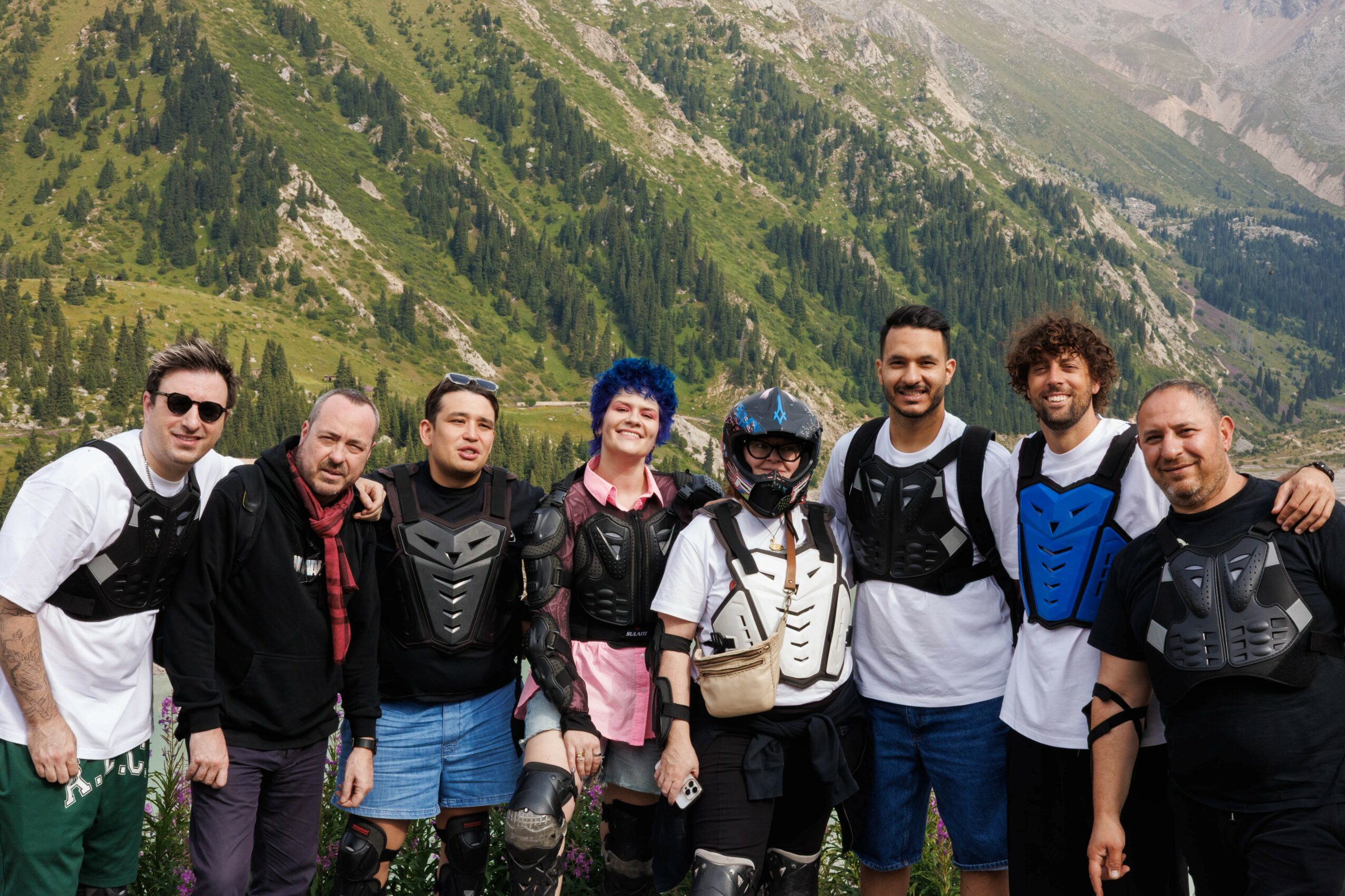 Group of eight people standing outdoors with mountains and trees in the background, smiling and posing for the photo.
