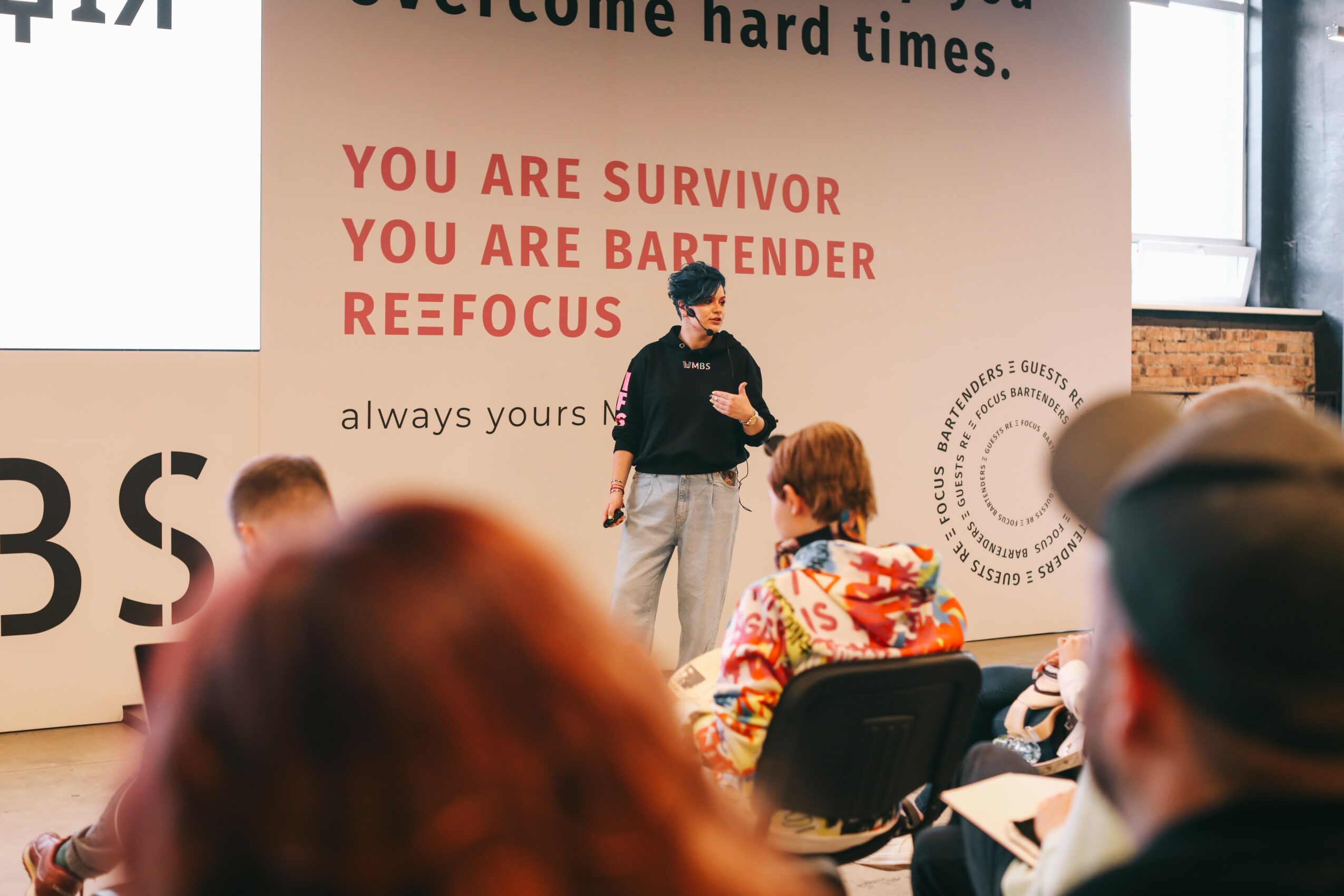 Person standing on stage speaking to seated audience in a room with a large screen and motivational text on the wall.