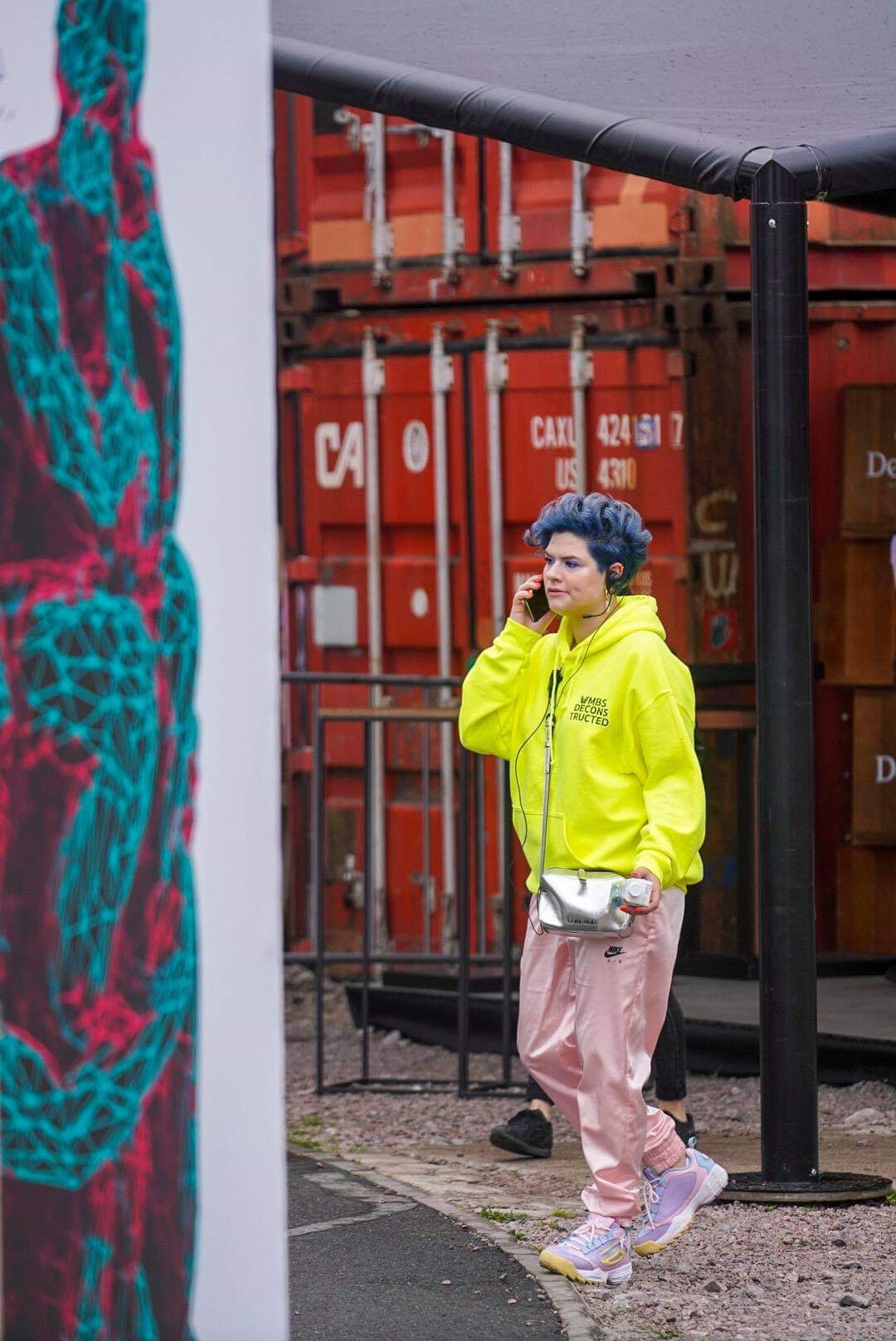 Person in bright yellow jacket and pink pants walking outdoors near shipping containers, holding a phone to their ear.