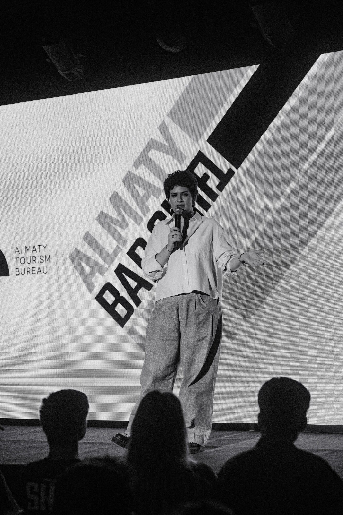Person speaking on stage with a large screen behind displaying text, audience silhouettes in foreground.