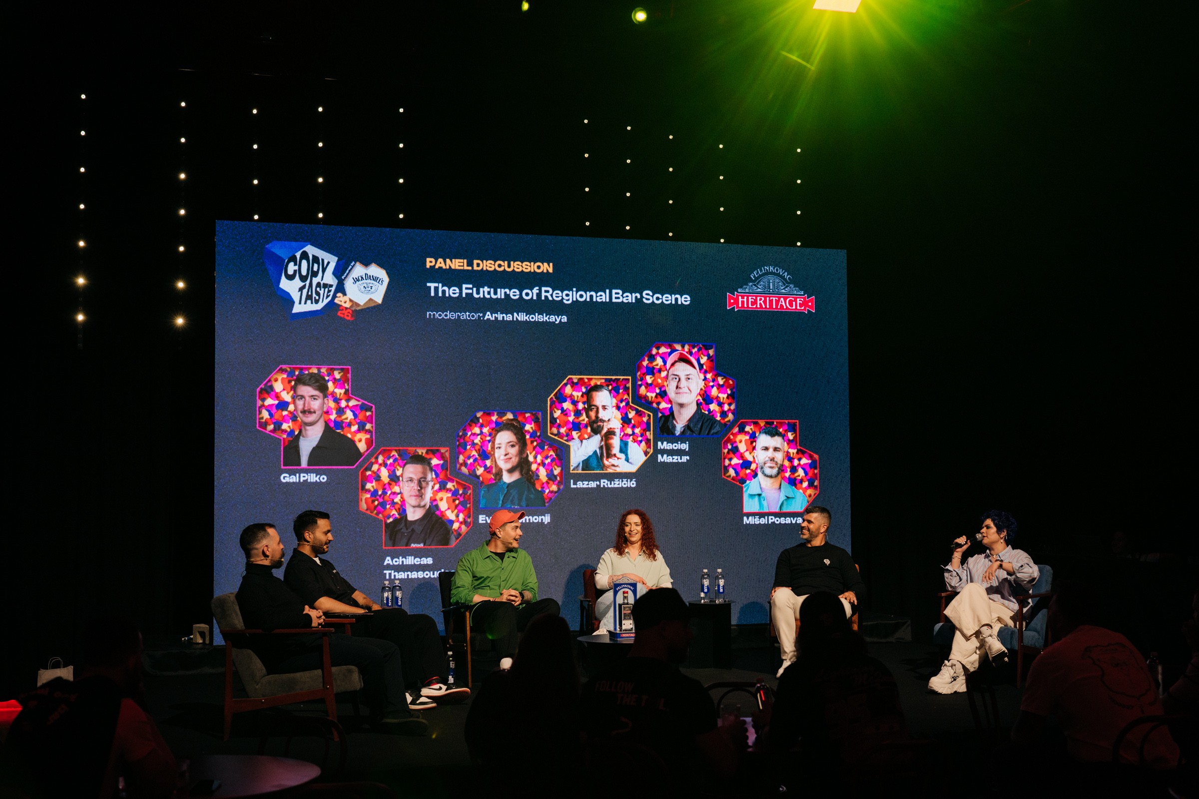 Panel discussion on stage with five people seated, large screen behind displaying images and text, green stage lighting.