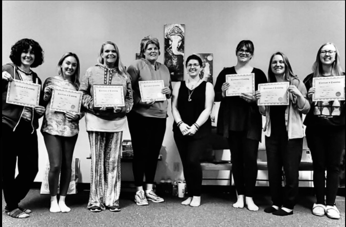 Group of nine people standing indoors, holding certificates, smiling, with a painting on the wall behind them.