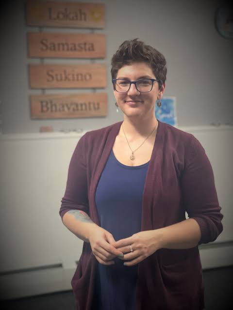 Woman with short curly hair and glasses smiling, standing indoors with a wall and signs in the background.
