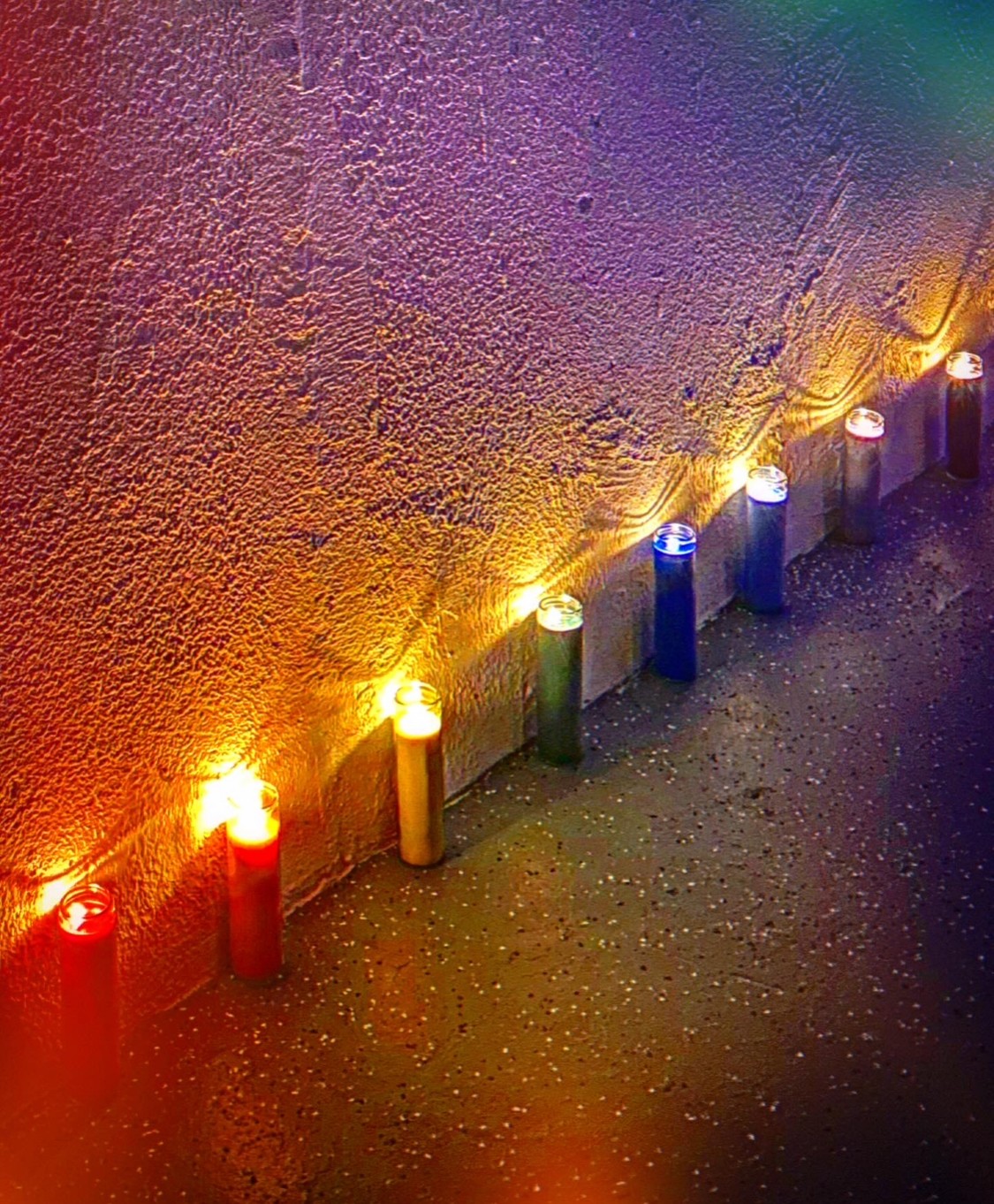 Colorful candles arranged in a row on a textured surface, illuminated against a wall with multicolored lighting.