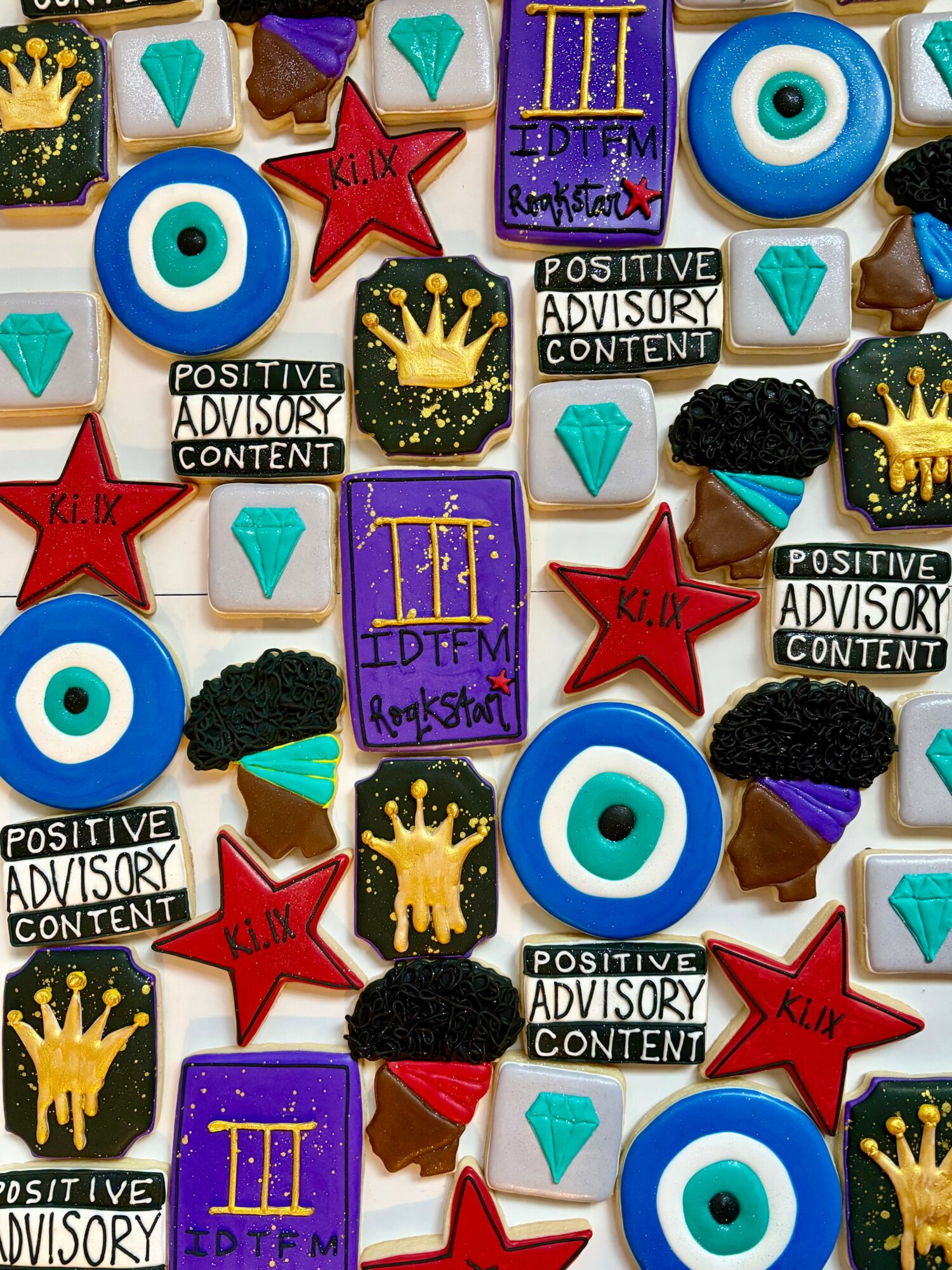 Assorted cookies with colorful icing and decorations, including eyes, crowns, stars, and text, arranged on a surface.