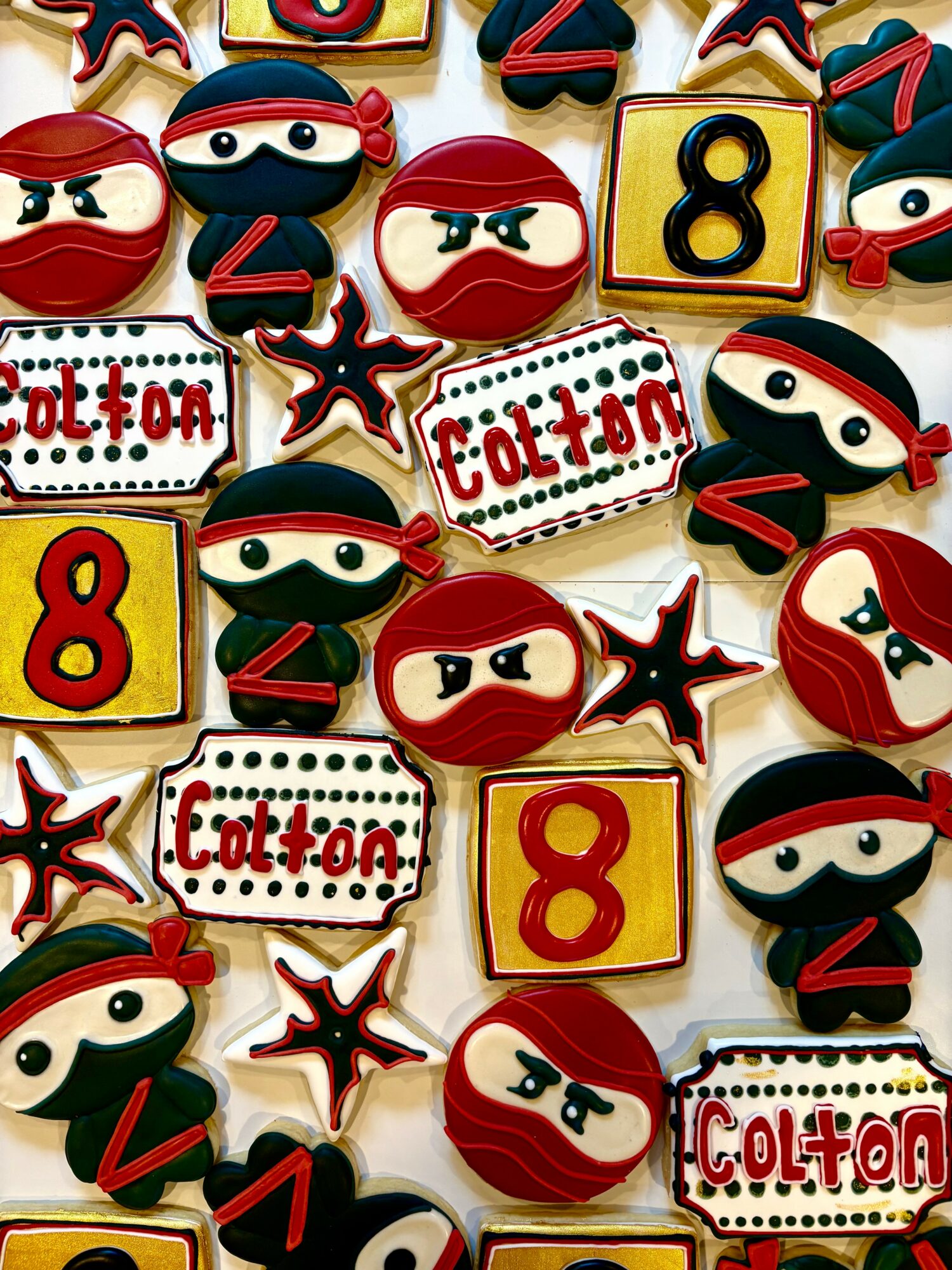 Assorted cookies with ninja, number 8, and 'Colton' designs, decorated in red, black, and yellow, on a white background.