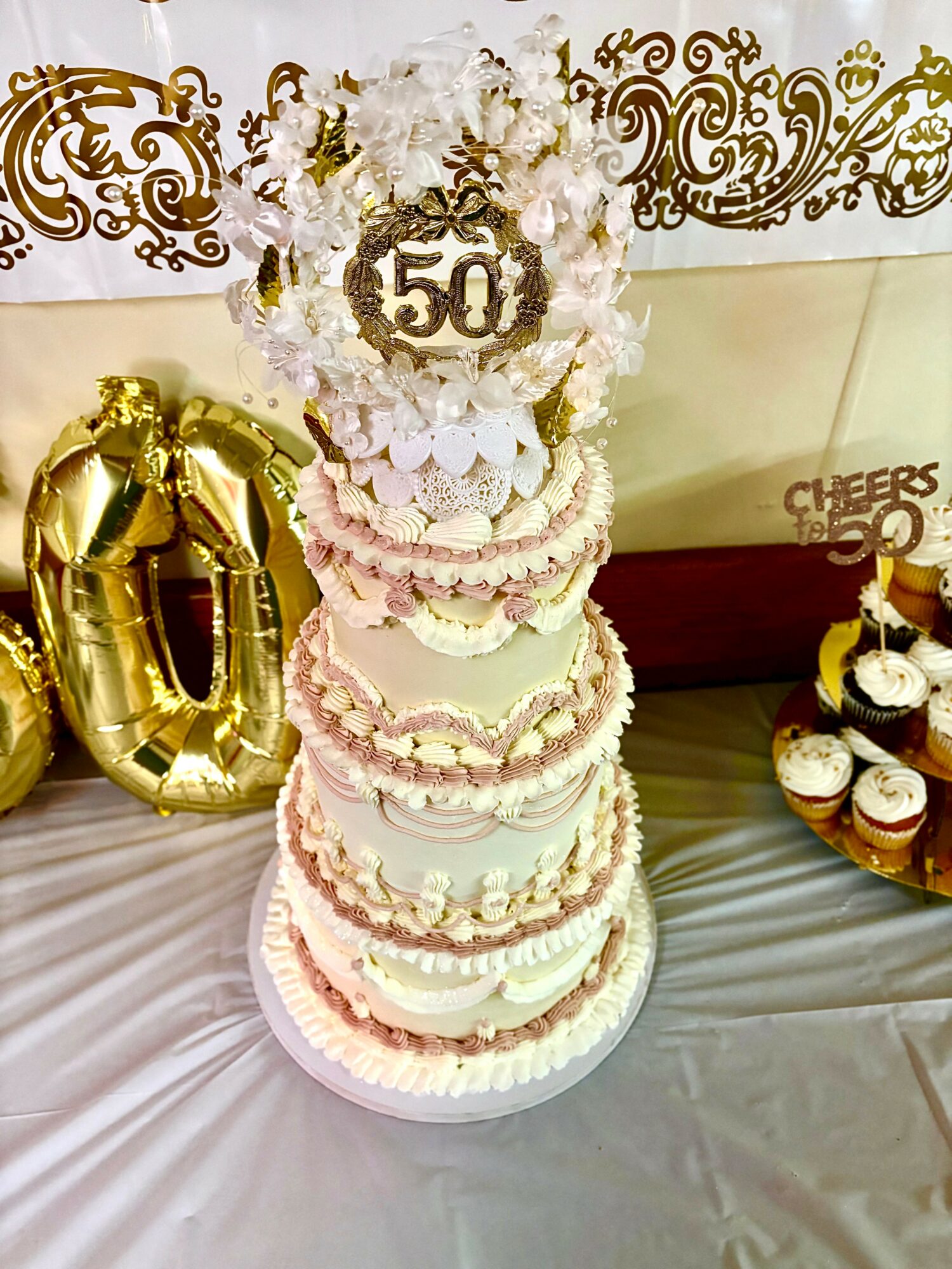 Multi-tiered cake with white and pink decorations, topped with a '50' sign and floral accents, celebrating a 50th anniversary.