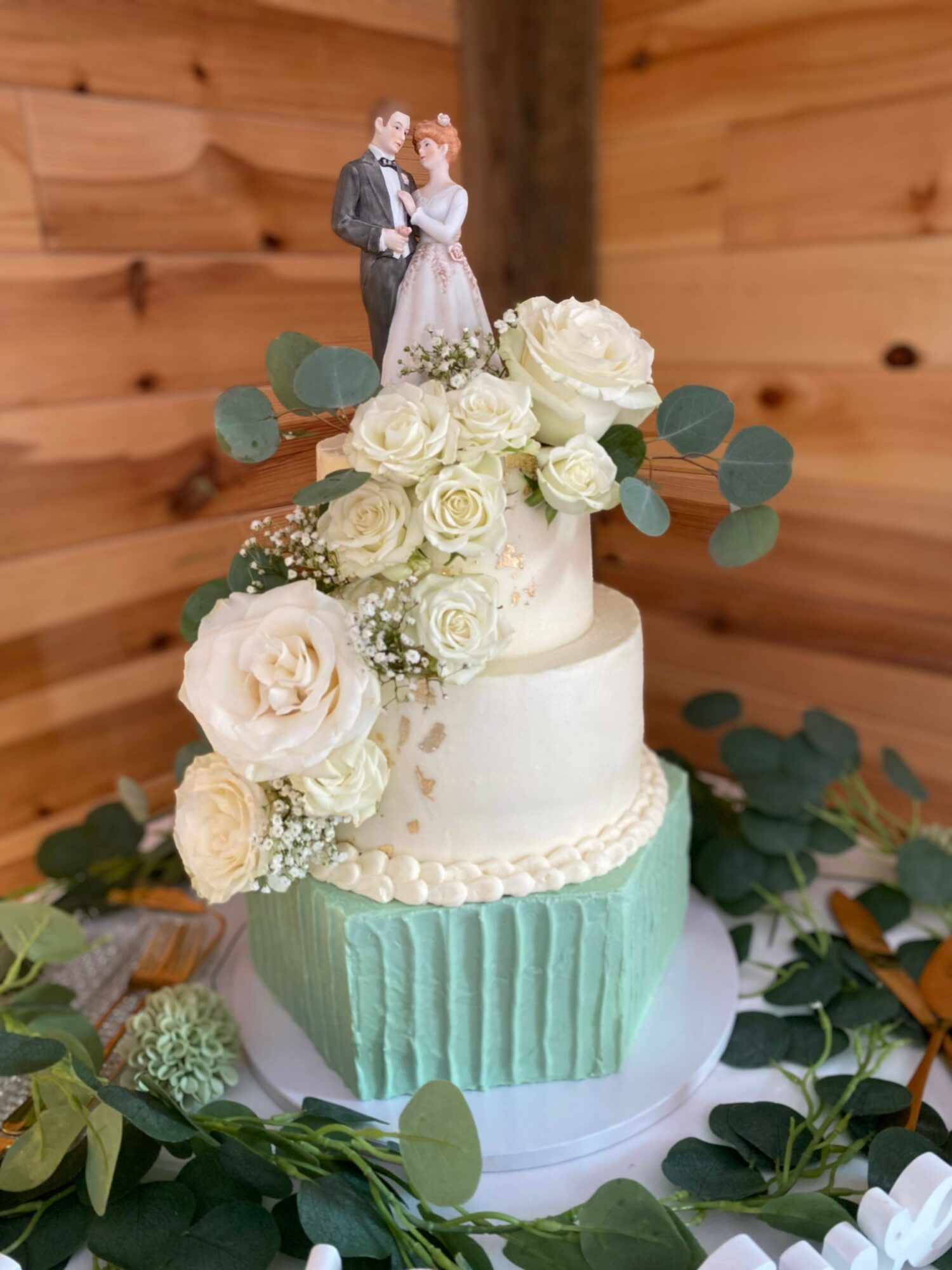 Three-tier wedding cake decorated with white roses, greenery, and a bride and groom figurine topper, set against a wooden background.
