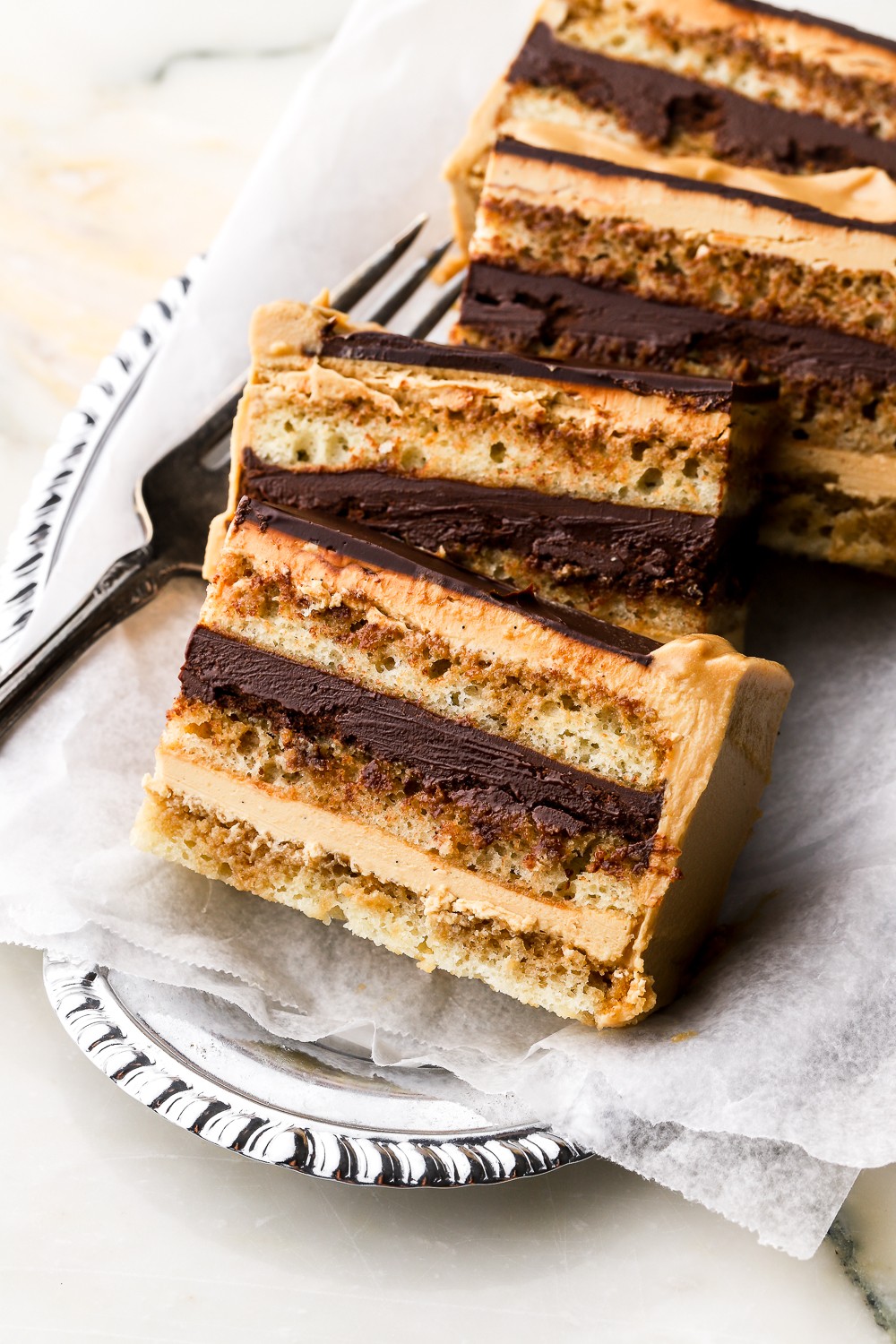 Two slices of layered cake with chocolate and cream, on a silver tray with parchment paper, fork nearby.