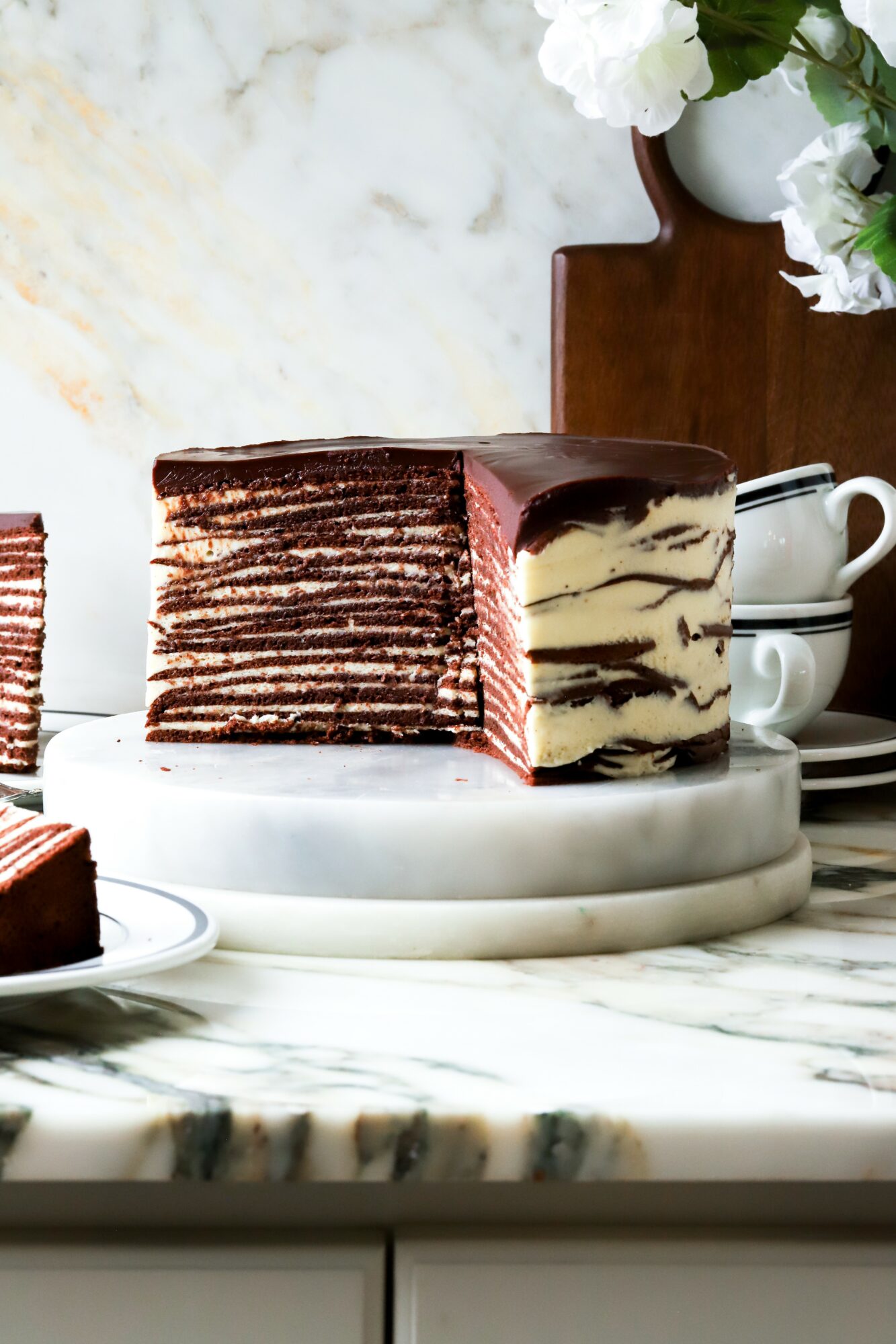 Layered chocolate cake with pink filling on a white marble surface, with a marble background and a wooden vase with white flowers in the background.