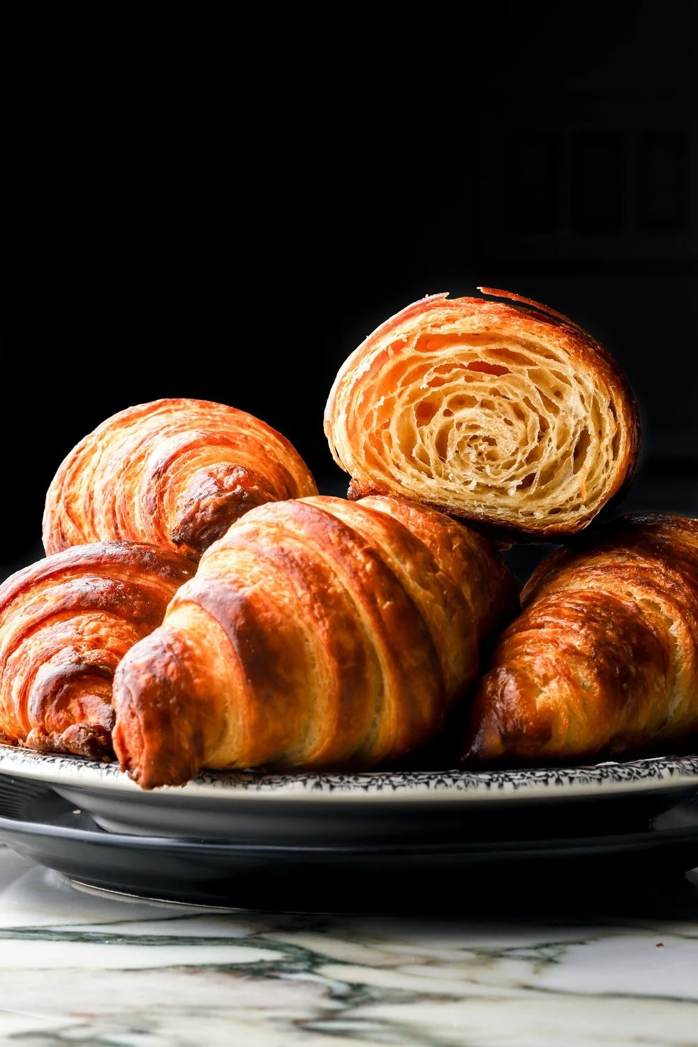 Several croissants on a plate, one cut open showing flaky layers, against a dark background.