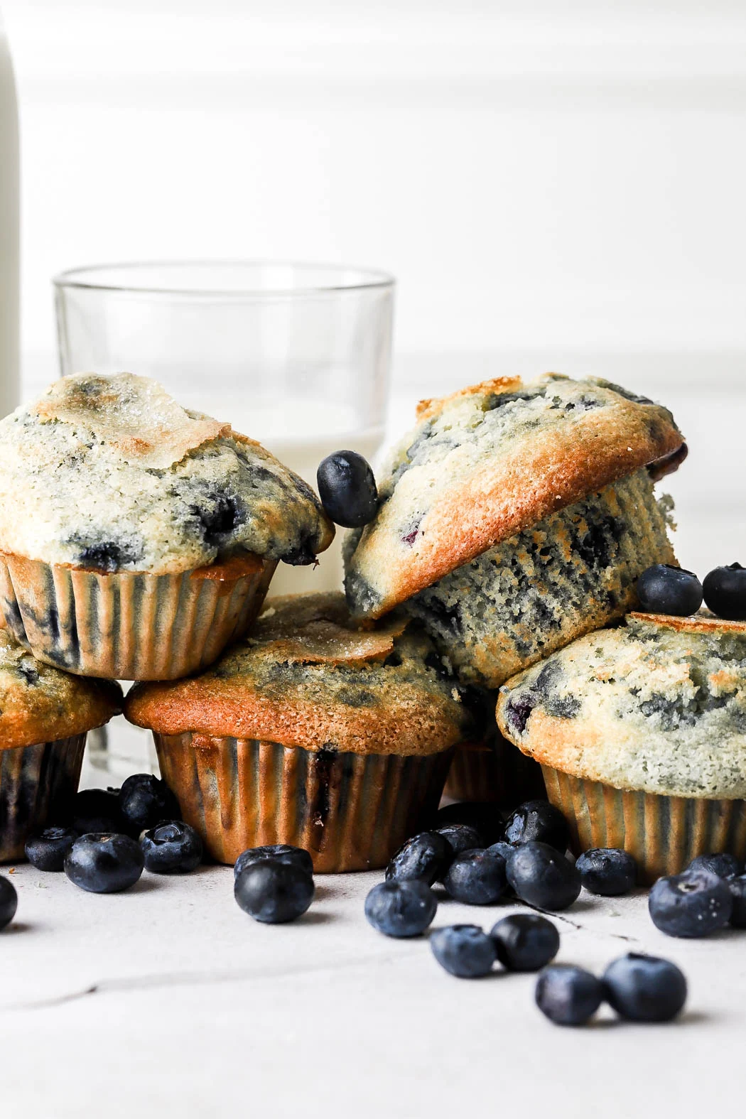 Blueberry muffins with blueberries scattered around, a glass of water in the background, on a white surface.