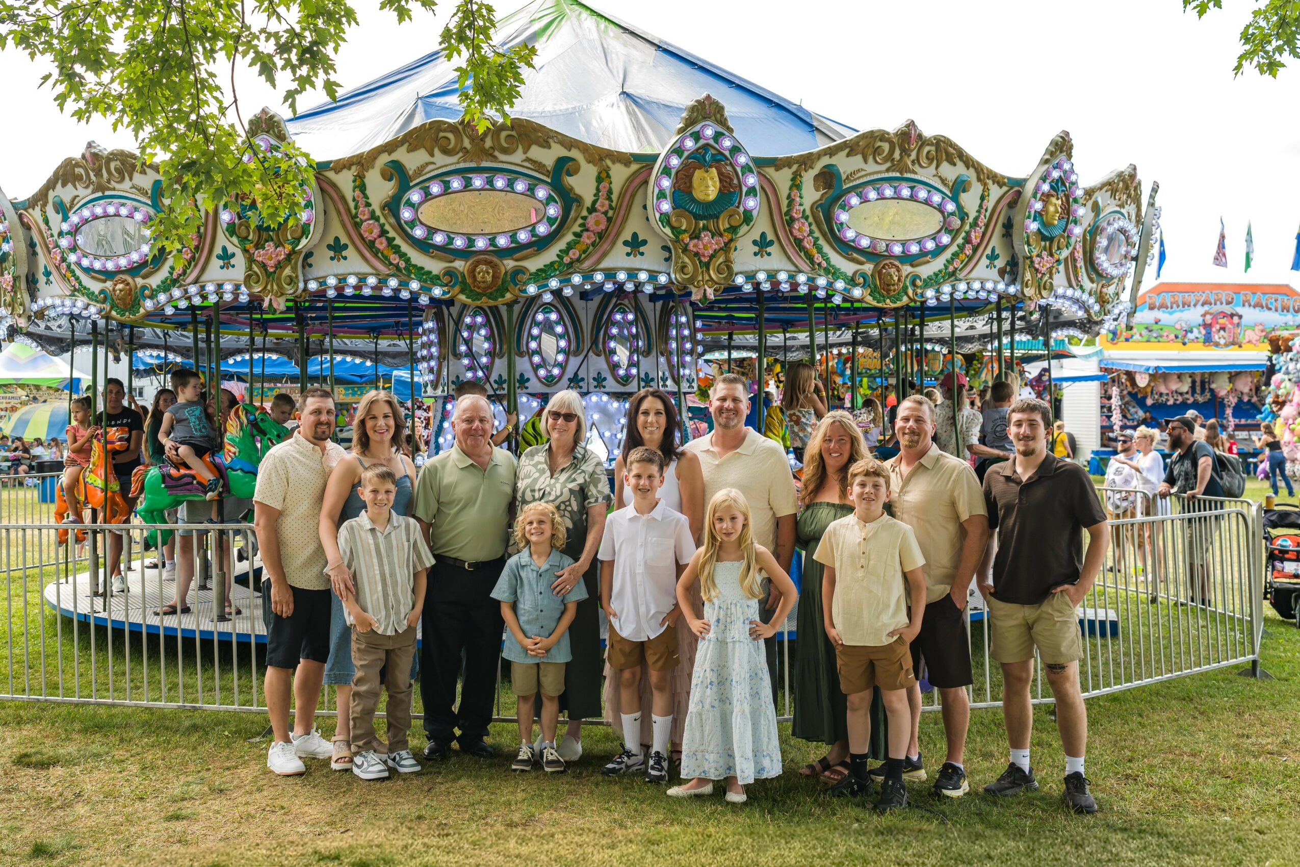 Group of people standing in front of a carousel at a fairground, with rides and booths in the background.