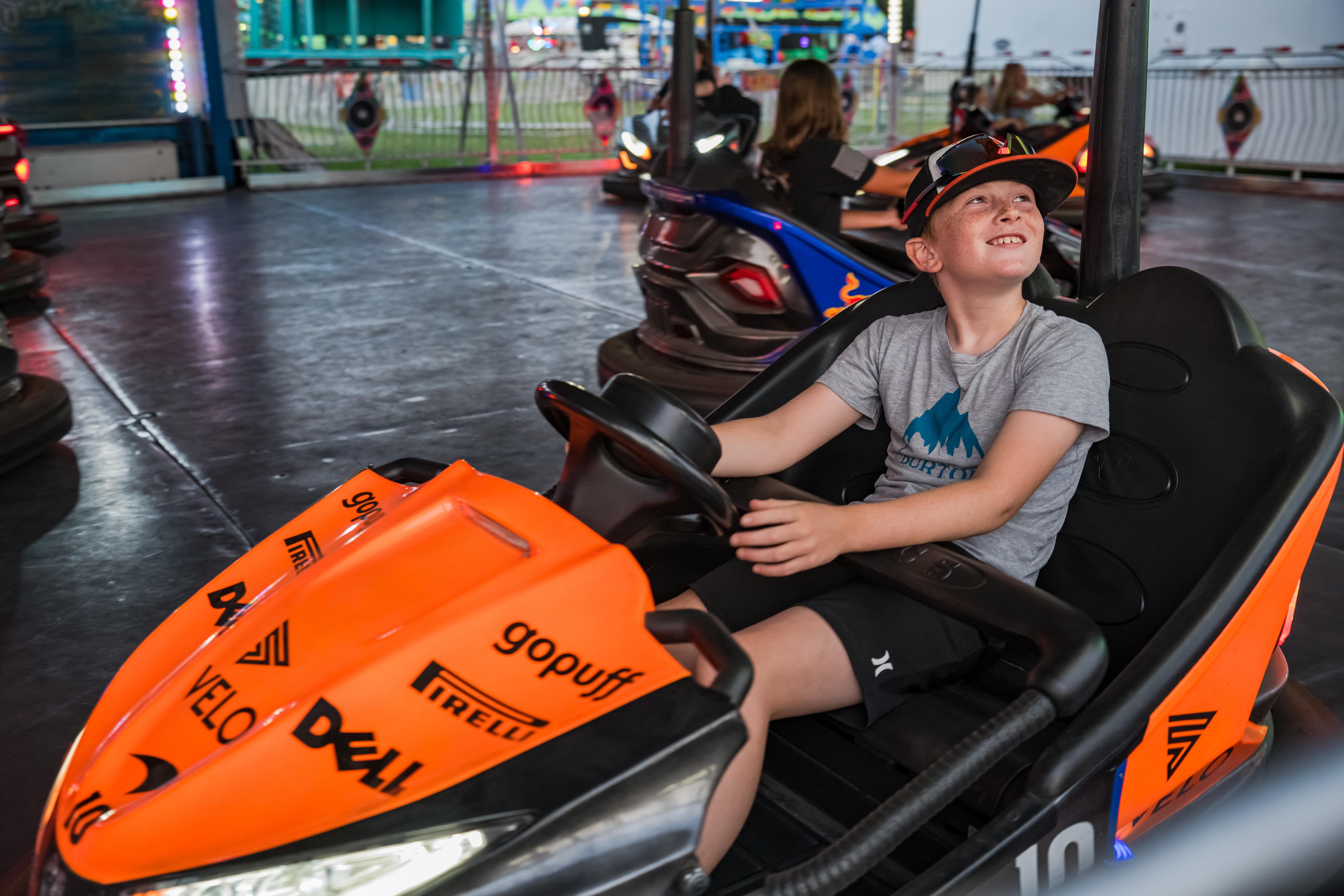 Child smiling and enjoying a ride in an orange go-kart at an amusement park, with other rides and people in the background.