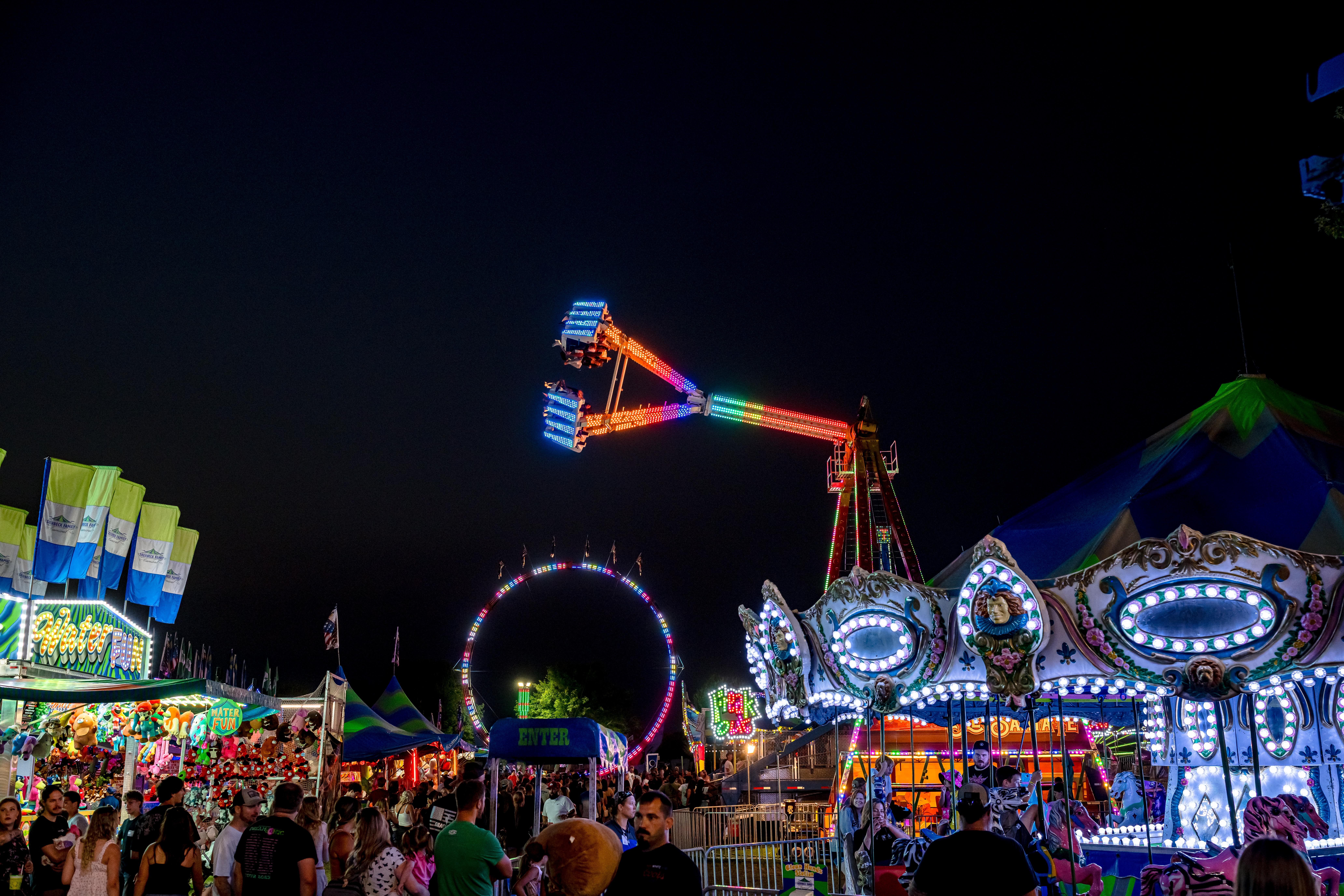 Nighttime carnival scene with illuminated rides, including a swinging ride, Ferris wheel, and carousel, with colorful lights and attractions.