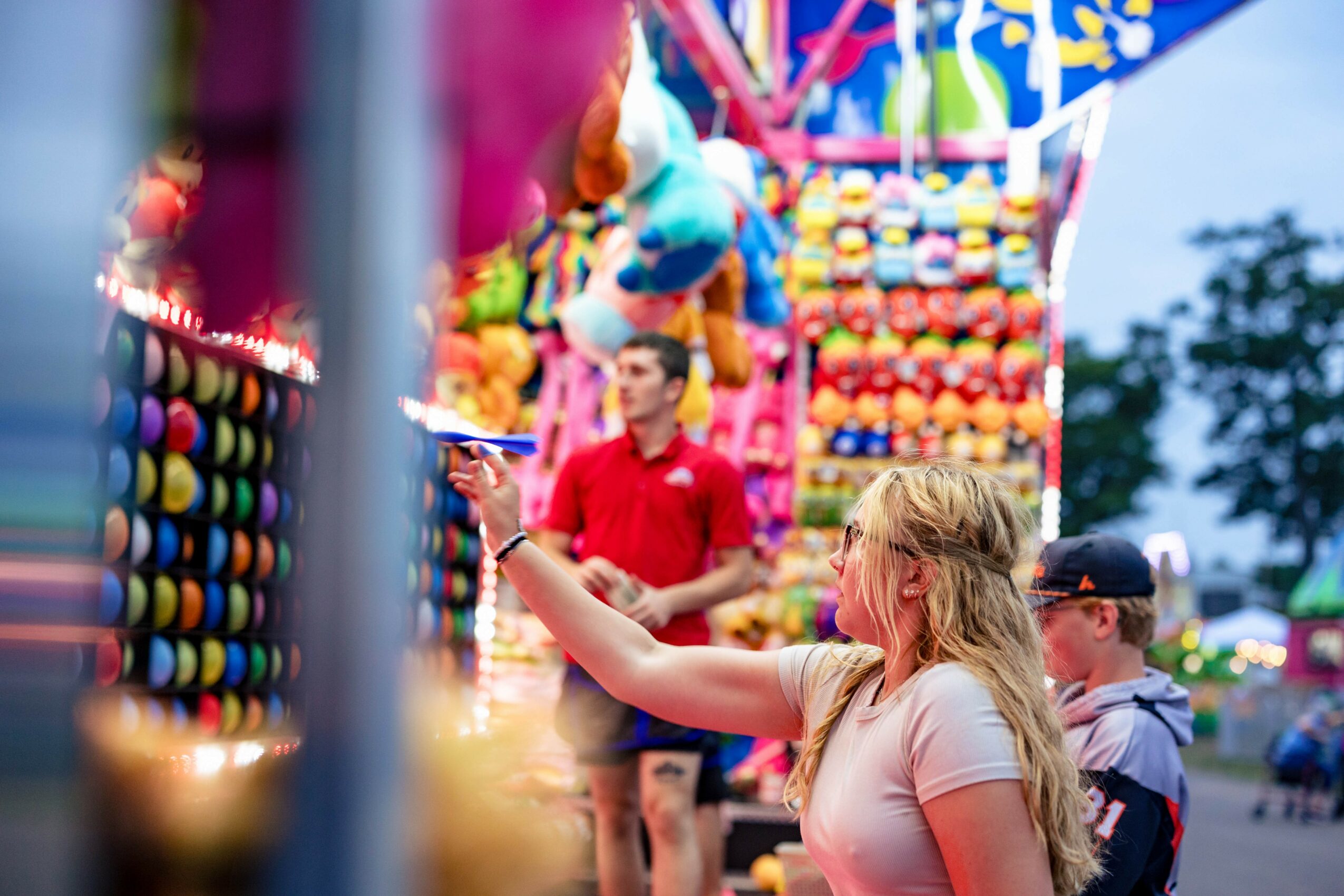 Girl with long blonde hair reaching for a prize at a colorful carnival game booth, with a man in a red shirt in the background.