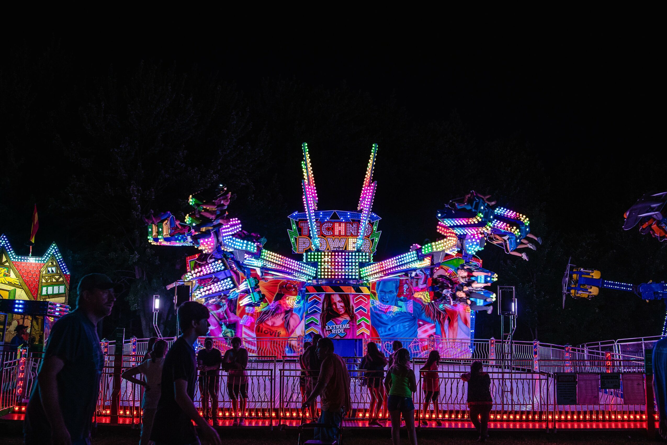People stand in front of a colorful amusement park ride illuminated with bright lights at night.