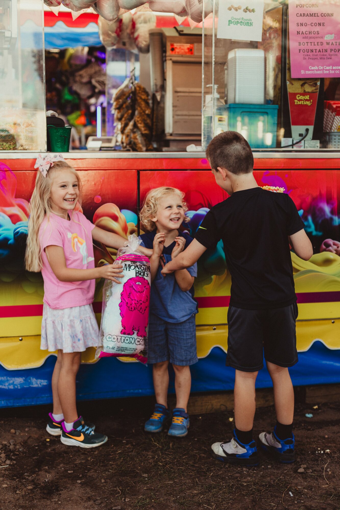 Three children, two girls and one boy, stand in front of a colorful food stand, smiling and talking.