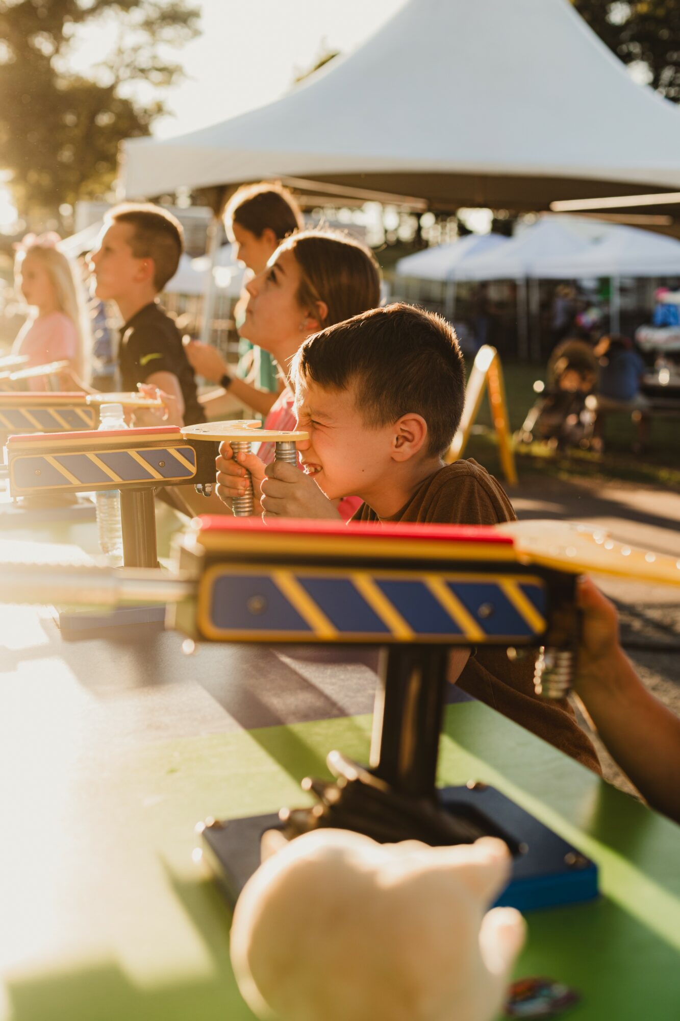 Children playing a game outdoors under sunlight, with tents and people in the background, during daytime.