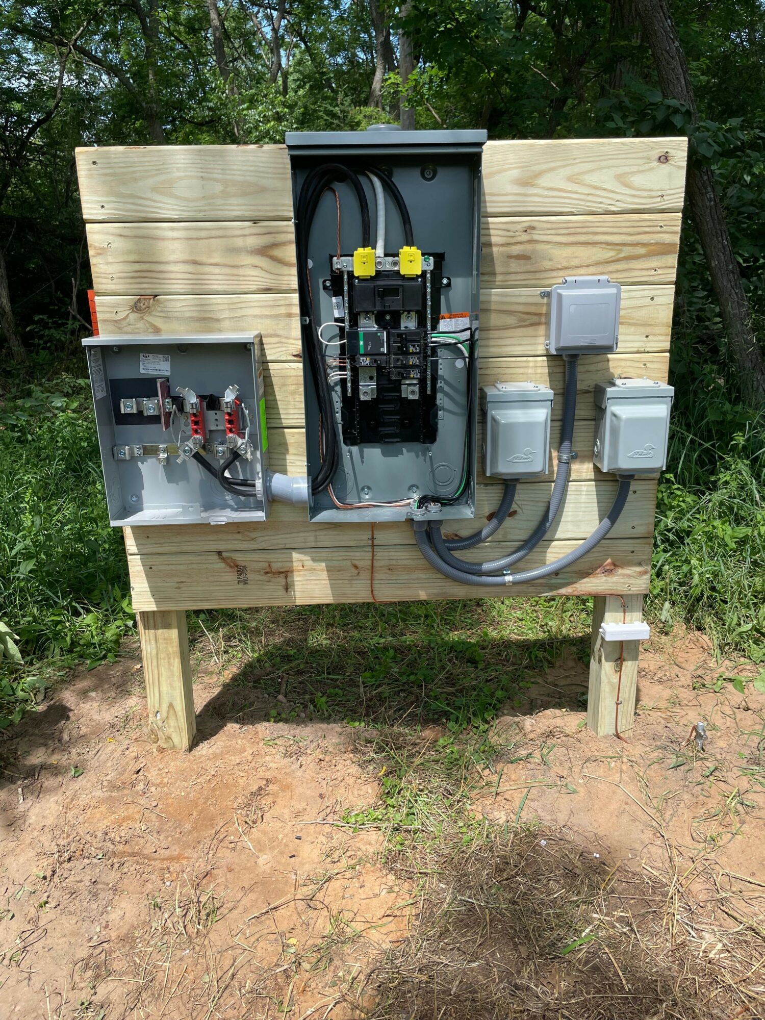 Electrical equipment mounted on a wooden panel outdoors, with wires and boxes, on a dirt and grass surface.