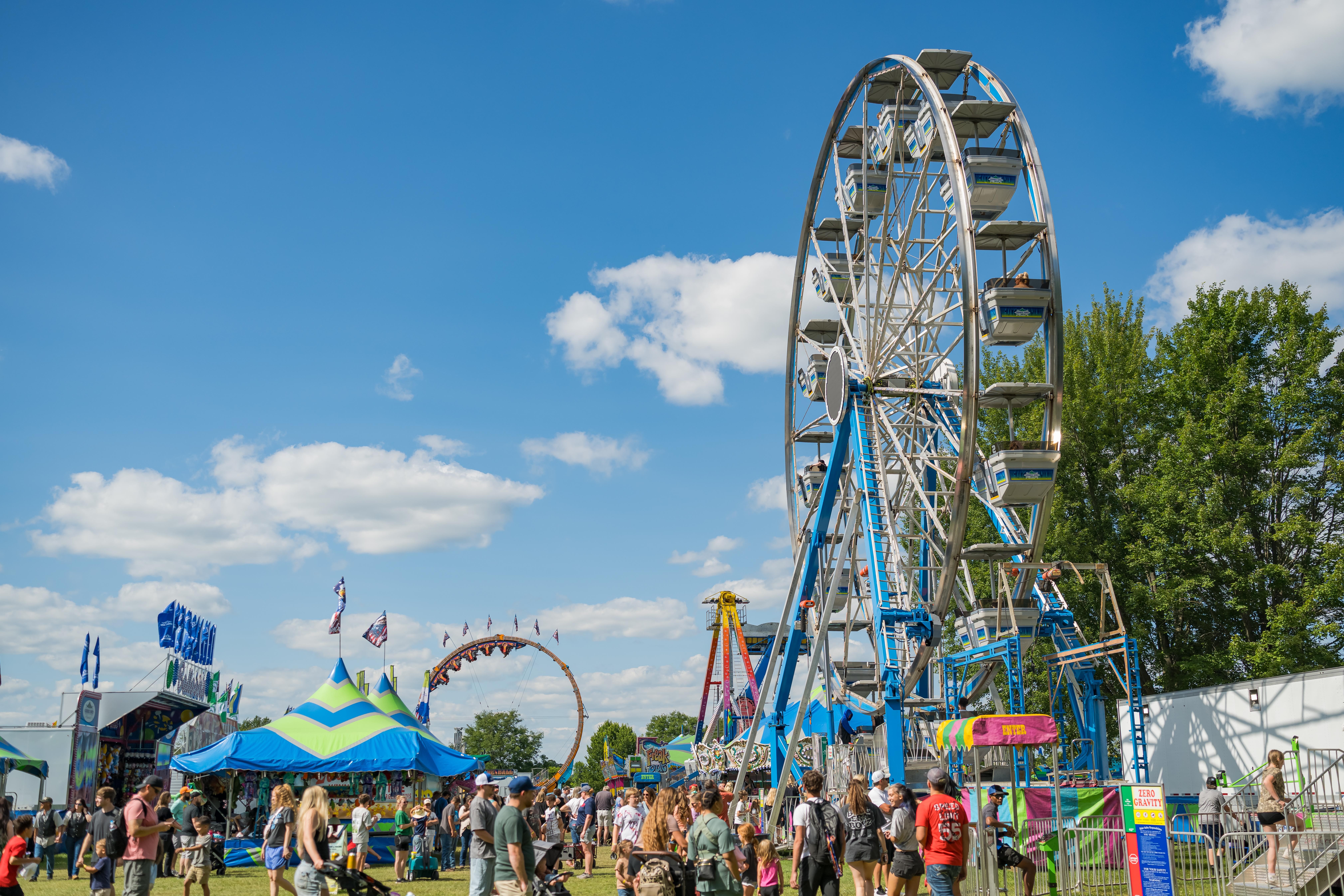 People at an amusement park with a large Ferris wheel, tents, and rides under a blue sky with clouds.