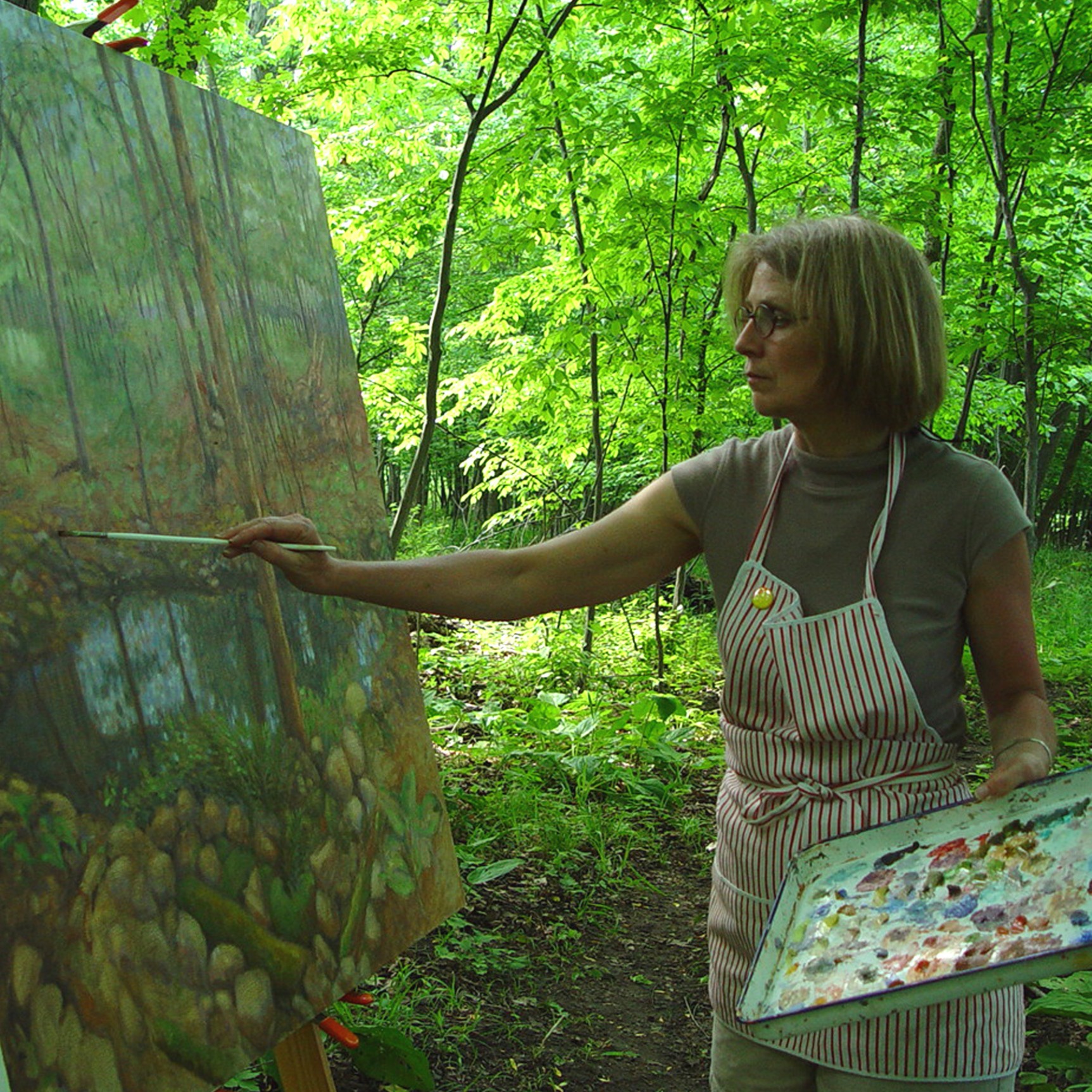 Woman painting outdoors in a green forest, wearing an apron and glasses, holding a paintbrush, with a large canvas.