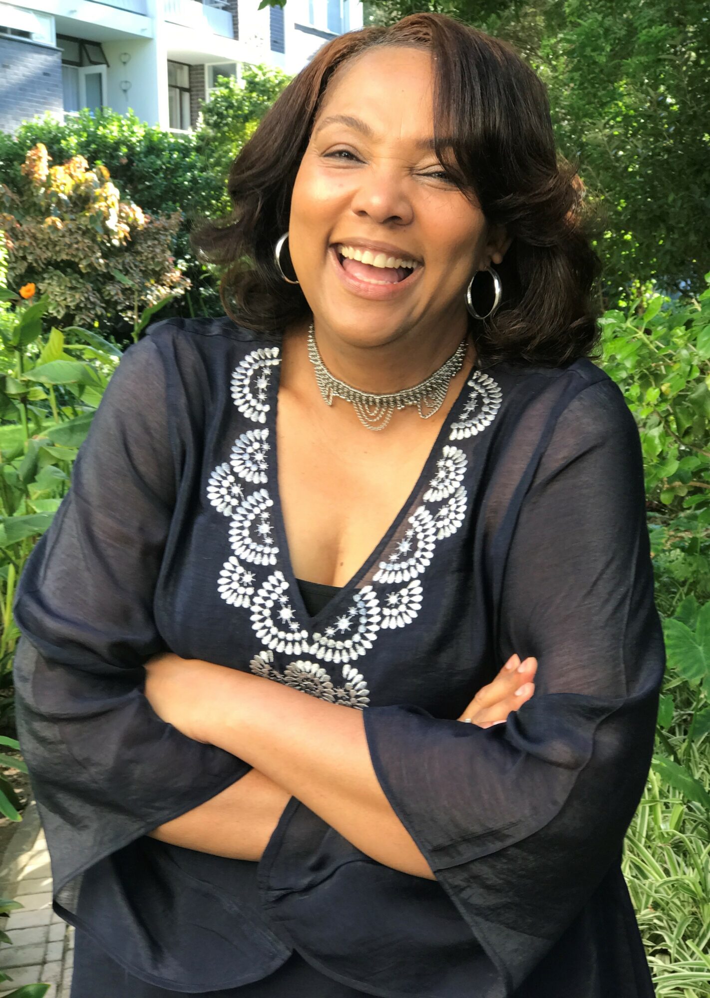 Woman smiling outdoors with arms crossed, wearing a dark blouse with white embroidery, surrounded by greenery.