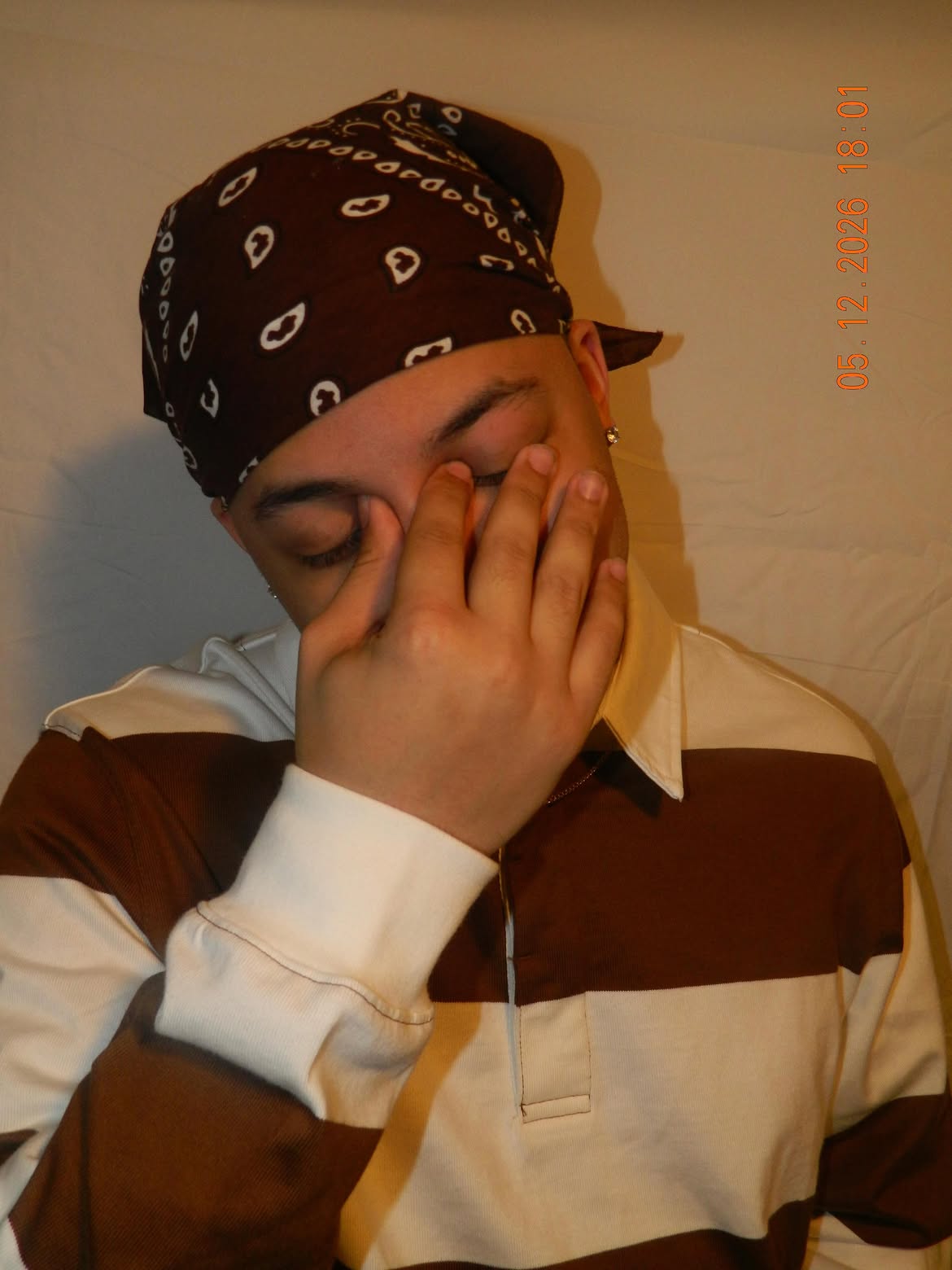 Person wearing a bandana and striped shirt, covering face with hand, against a plain background.
