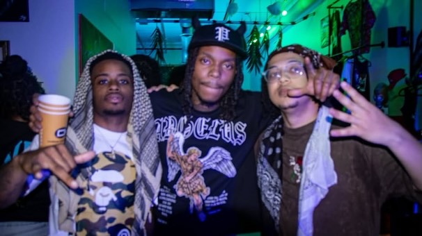 Three people posing together in a colorful indoor setting, one holding a cup, all making gestures, with vibrant lighting.